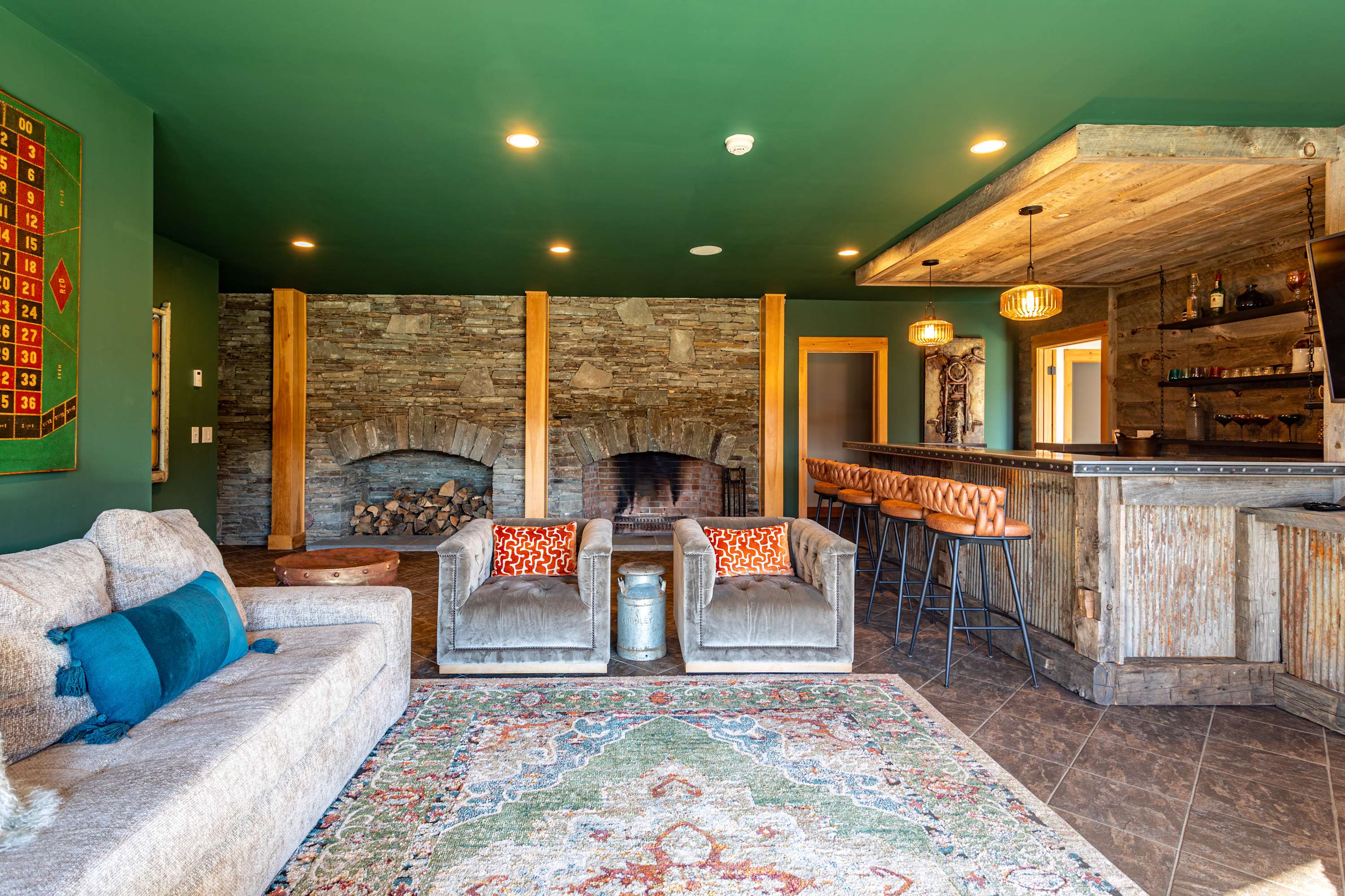 A spacious living room with a large sectional sofa and two armchairs facing a grand stone fireplace, with a colorful area rug defining the space. The rustic bar area is visible in the background, all under a vibrant green ceiling.
