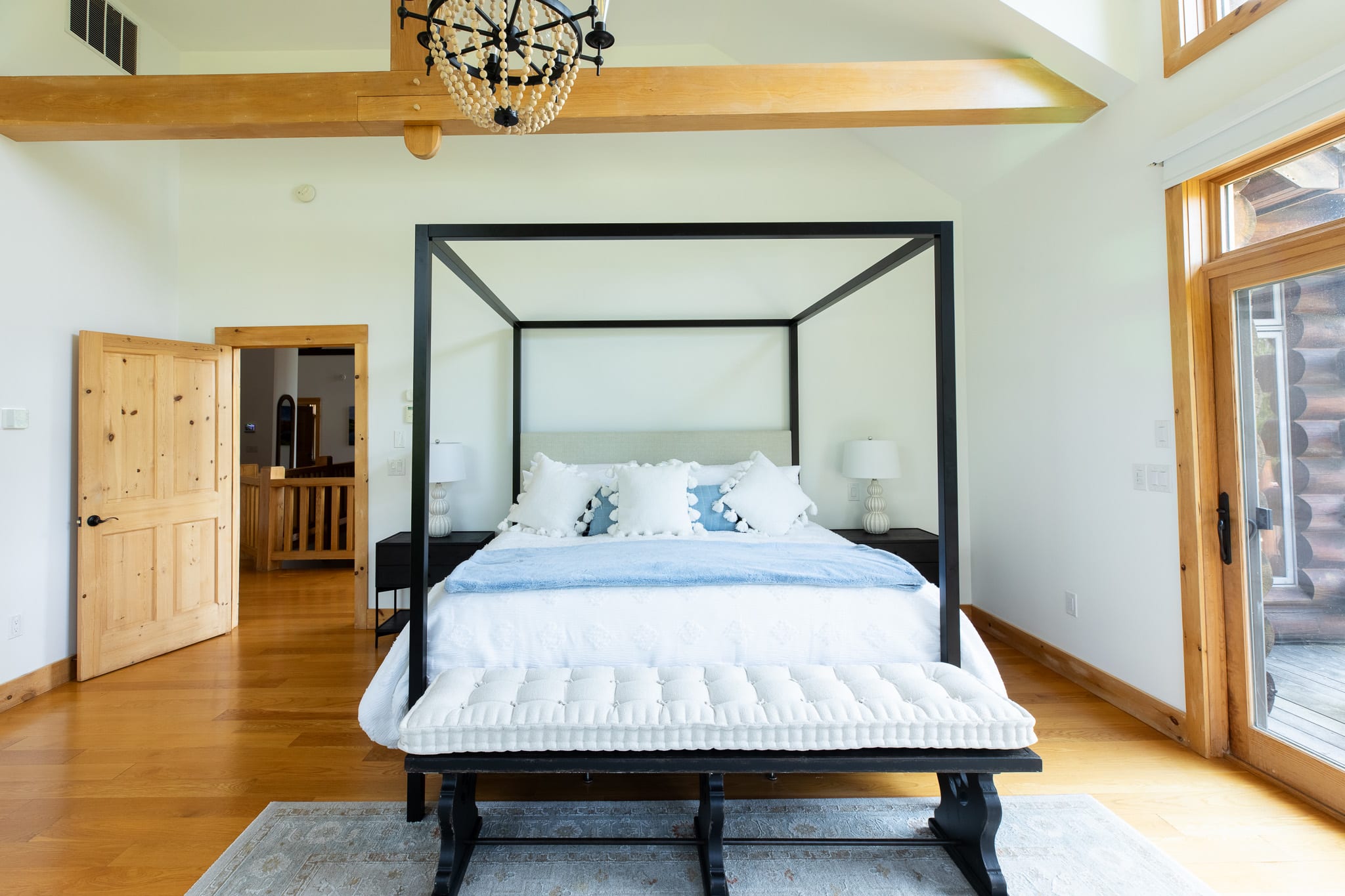 A close-up view of the black four-poster canopy bed with white and blue bedding, centered on a light-colored area rug. The room features warm hardwood floors, vaulted ceilings with wooden beams, and a wooden door leading to another room.