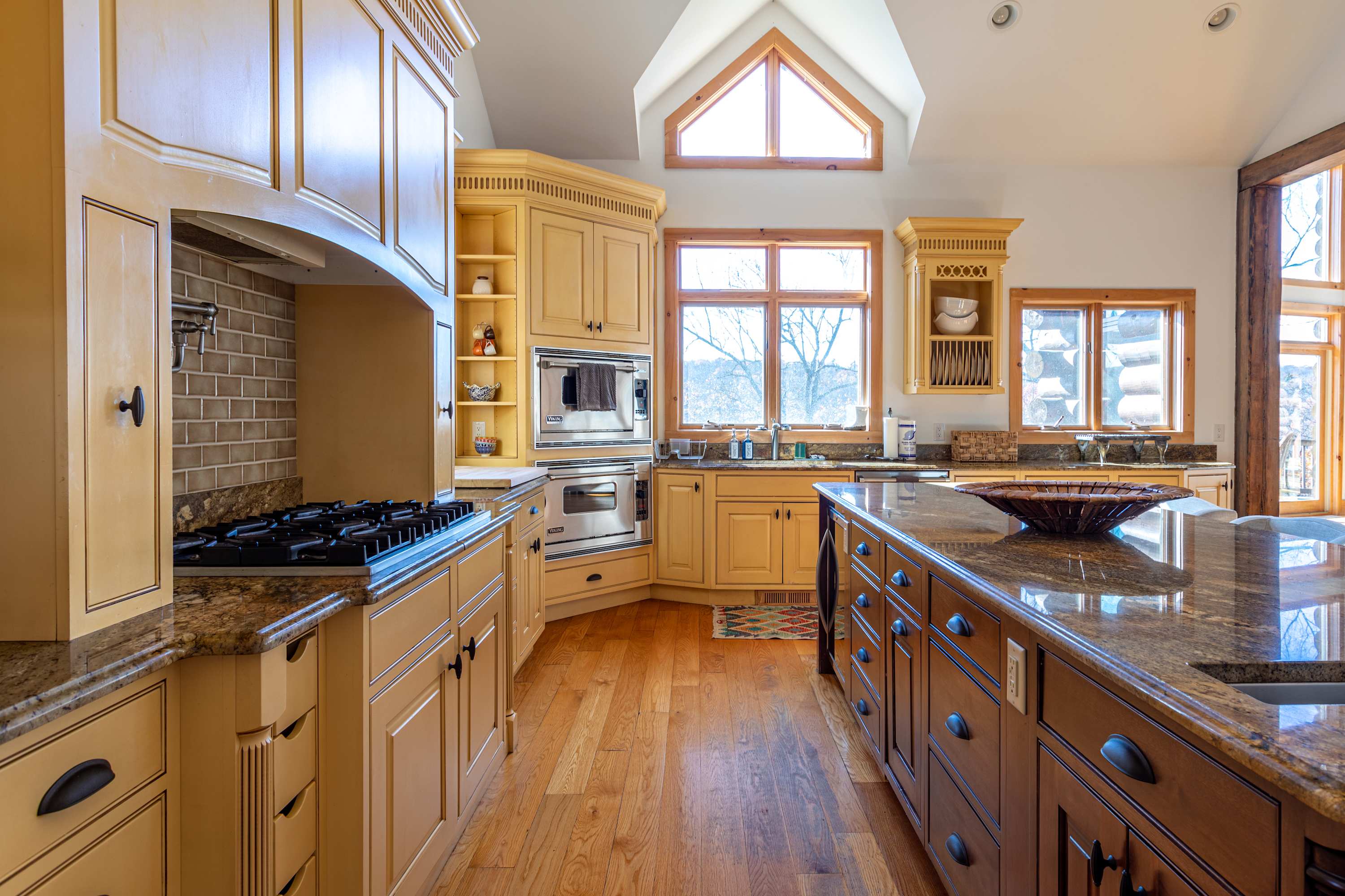 Corner view of the kitchen showing light yellow cabinets, a double oven, a sink under a window, and a large wooden island with a granite countertop, all on hardwood floors.