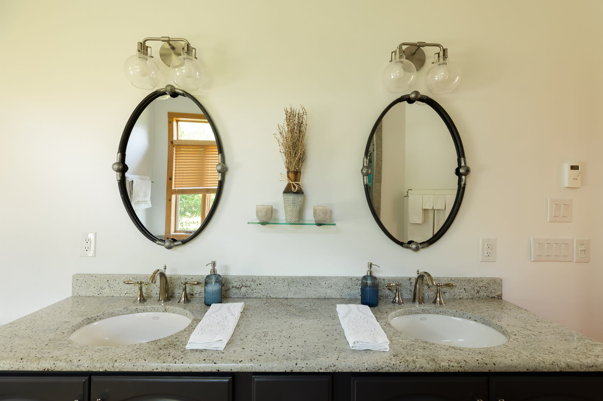 A close-up of the double vanity in the bathroom, showcasing the light granite countertop, two undermount sinks with brushed nickel faucets, and oval mirrors with modern sconce lights. A decorative vase sits between the mirrors.