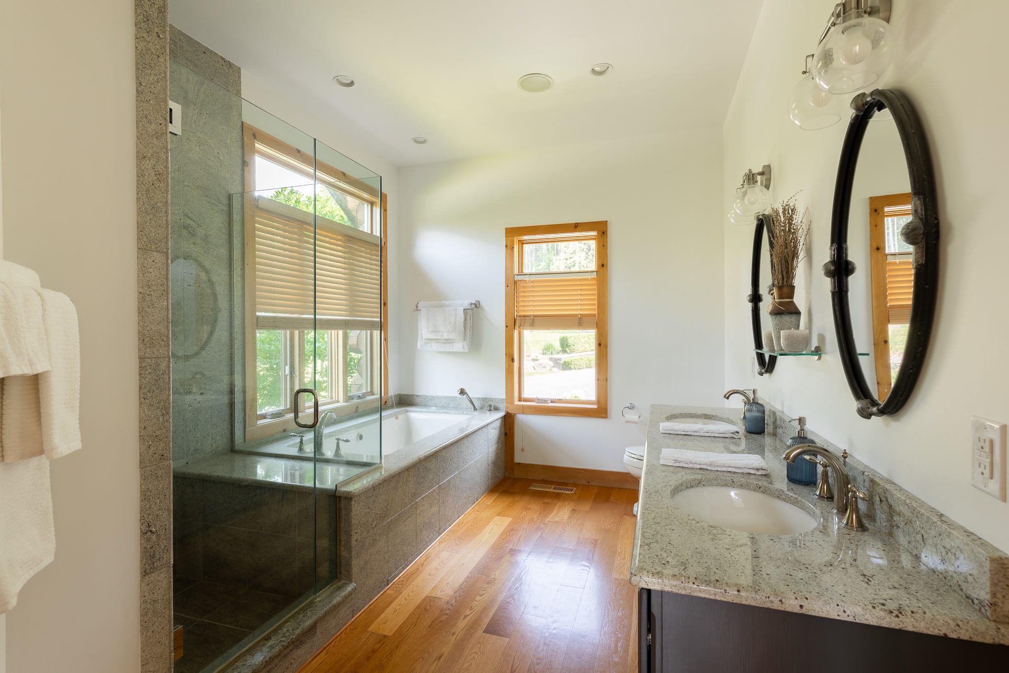 A spacious bathroom with a double vanity featuring a light granite countertop, two undermount sinks, and dark wood cabinets. Two oval mirrors with sconce lighting hang above, and a jetted tub and walk-in shower are visible.