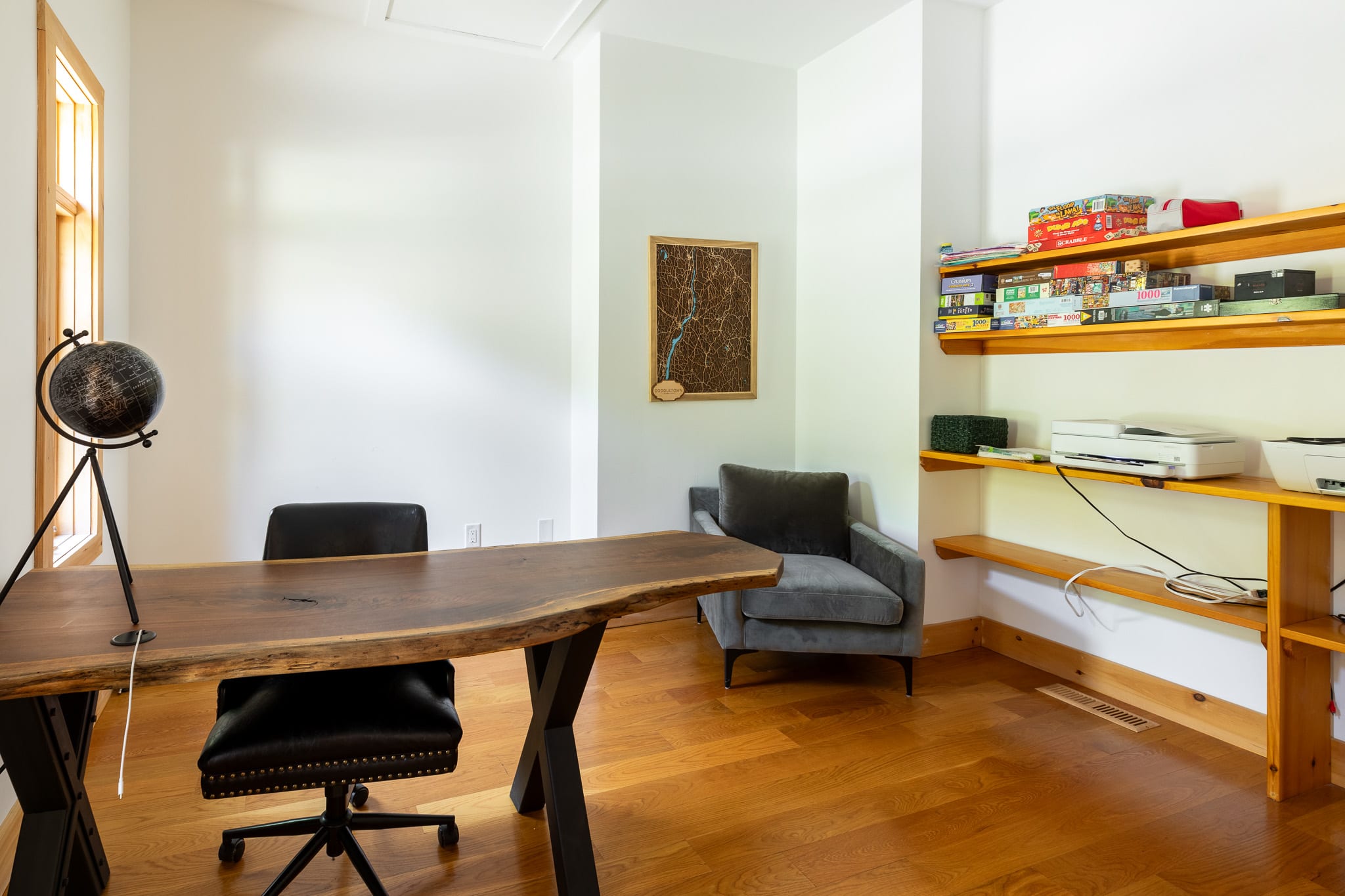 A bright office space with white walls and warm hardwood floors, featuring a live-edge wooden desk with a black leather chair. Built-in wooden shelves on the right hold board games, and a gray armchair sits in the corner.