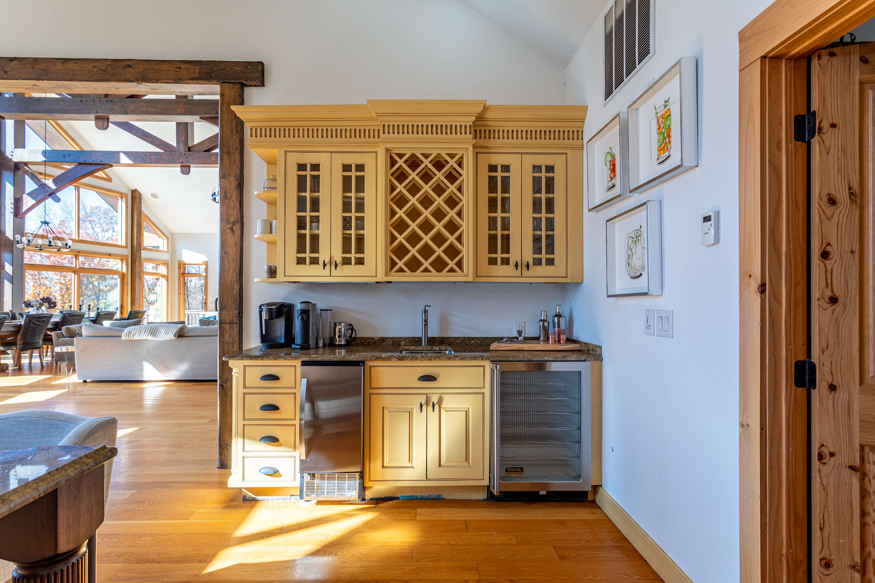 A bright wet bar area features light yellow cabinetry, a granite countertop with a sink, a mini-fridge, and a wine rack, all set on warm hardwood floors. Artwork adorns the white wall, and a large living area is visible in the background.