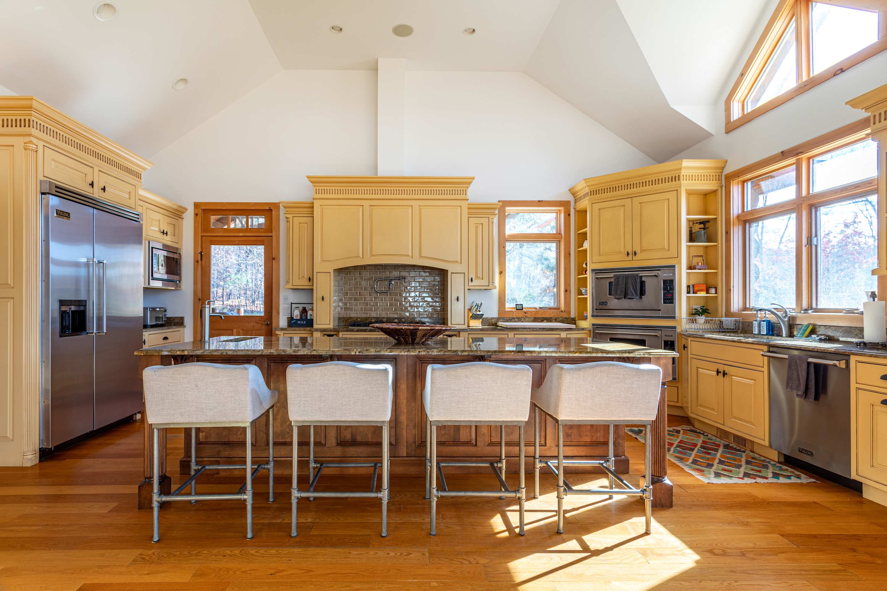 Spacious kitchen featuring light yellow cabinetry, a large wooden island with four white bar stools, stainless steel appliances, and vaulted ceilings with recessed lighting.