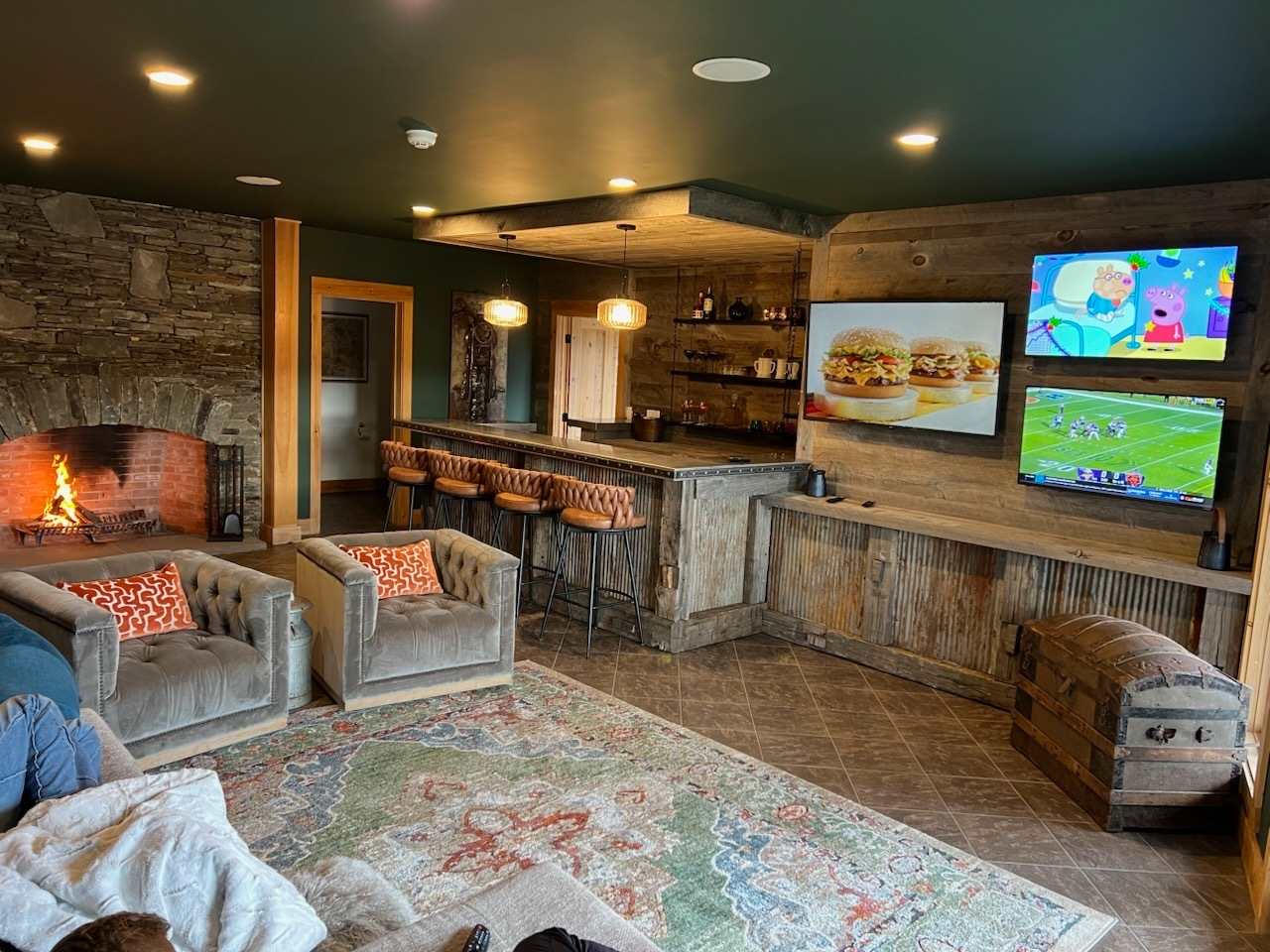 A lively entertainment space featuring a rustic bar with multiple bar stools, a comfortable seating area with sofas and armchairs, and a wall-mounted array of televisions. A large stone fireplace adds a cozy element to the room with its vibrant green ceiling.