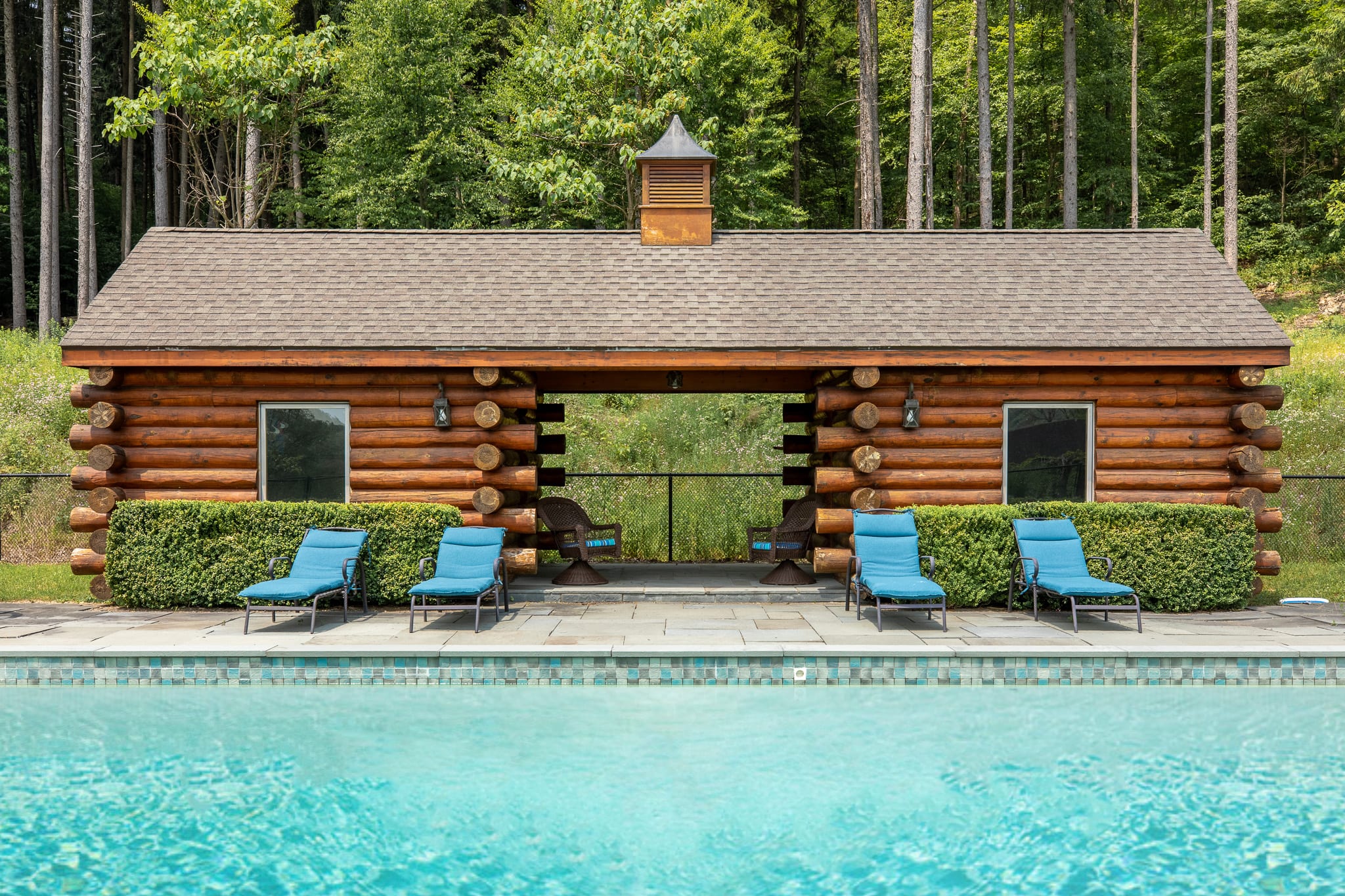 A clear blue swimming pool in the foreground, with a rustic log cabin pool house featuring a shingled roof and a central cupola, backed by dense green forest.