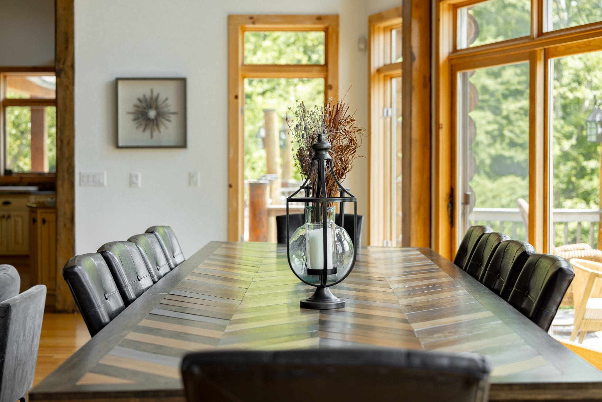 A detailed view of the rustic wooden dining table centerpiece, featuring a glass vase with dried botanicals and two black lanterns, highlighting the table's herringbone pattern and the dark leather chairs.