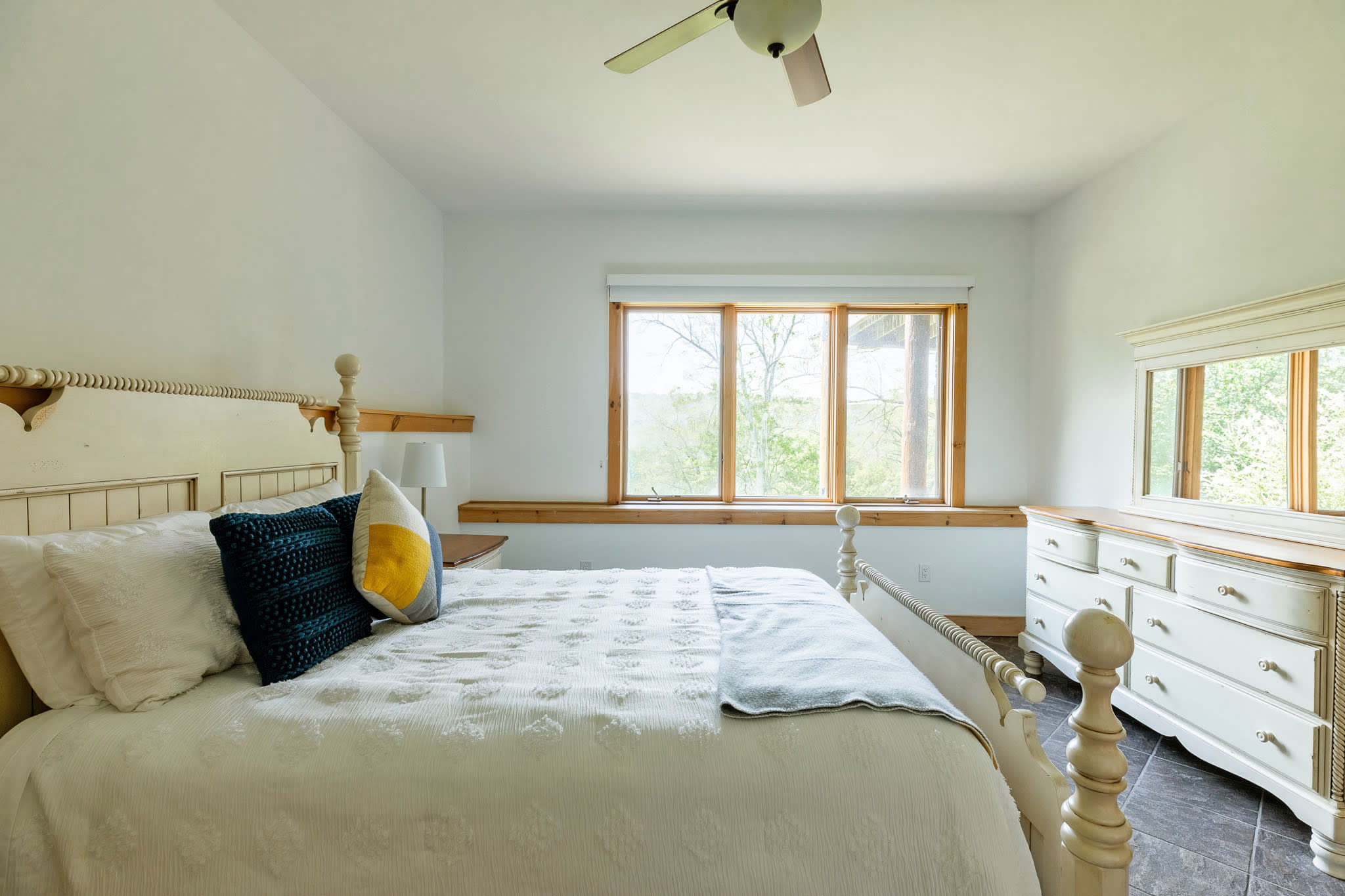 A wide shot of a bright bedroom with a white wooden queen bed, white bedding, and colorful throw pillows, featuring large windows with natural wood trim and a white dresser with a mirror.