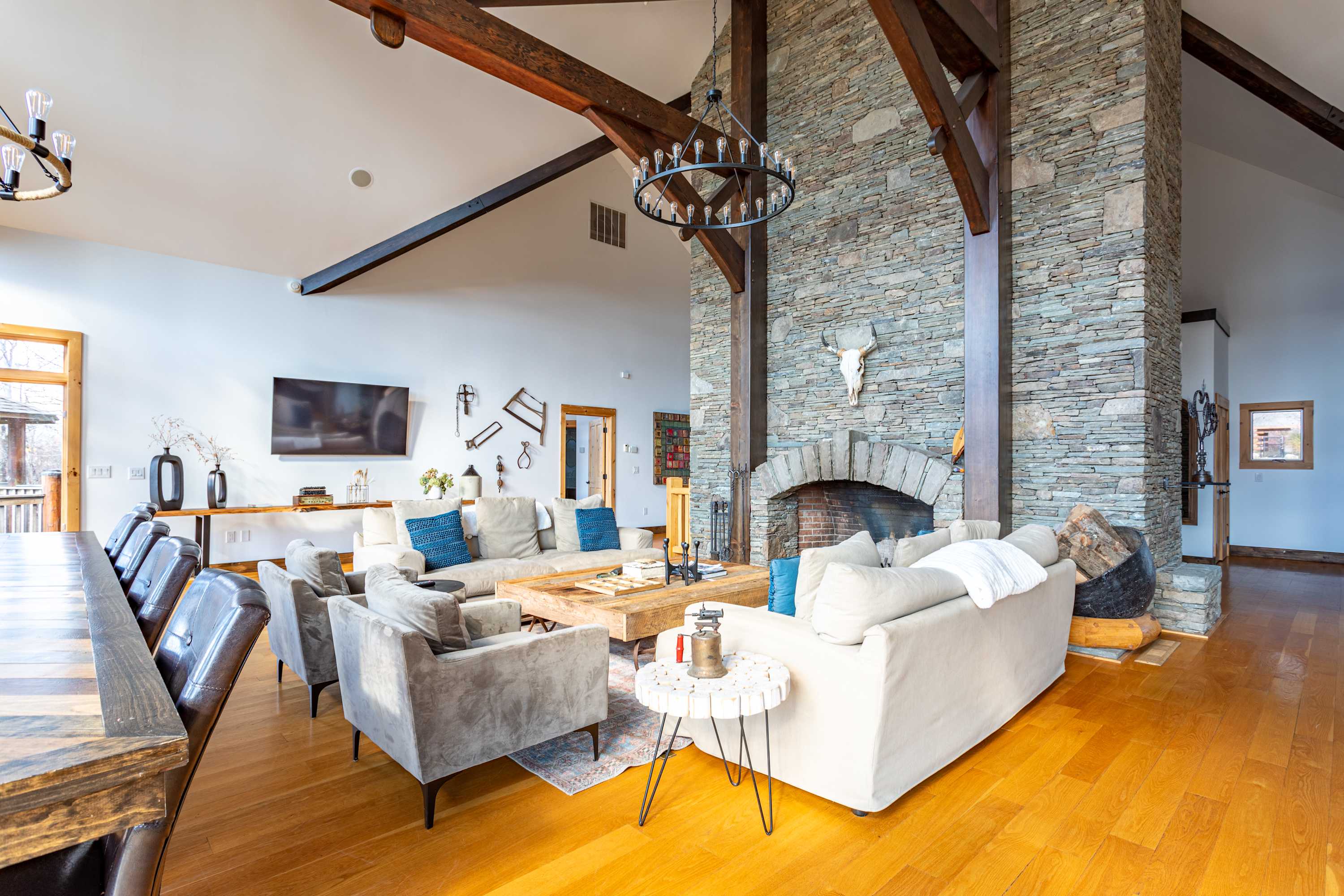 A spacious living room with multiple seating options including sofas and armchairs, a large wooden coffee table, and a wall-mounted TV, all anchored by a prominent stone fireplace and exposed beams.