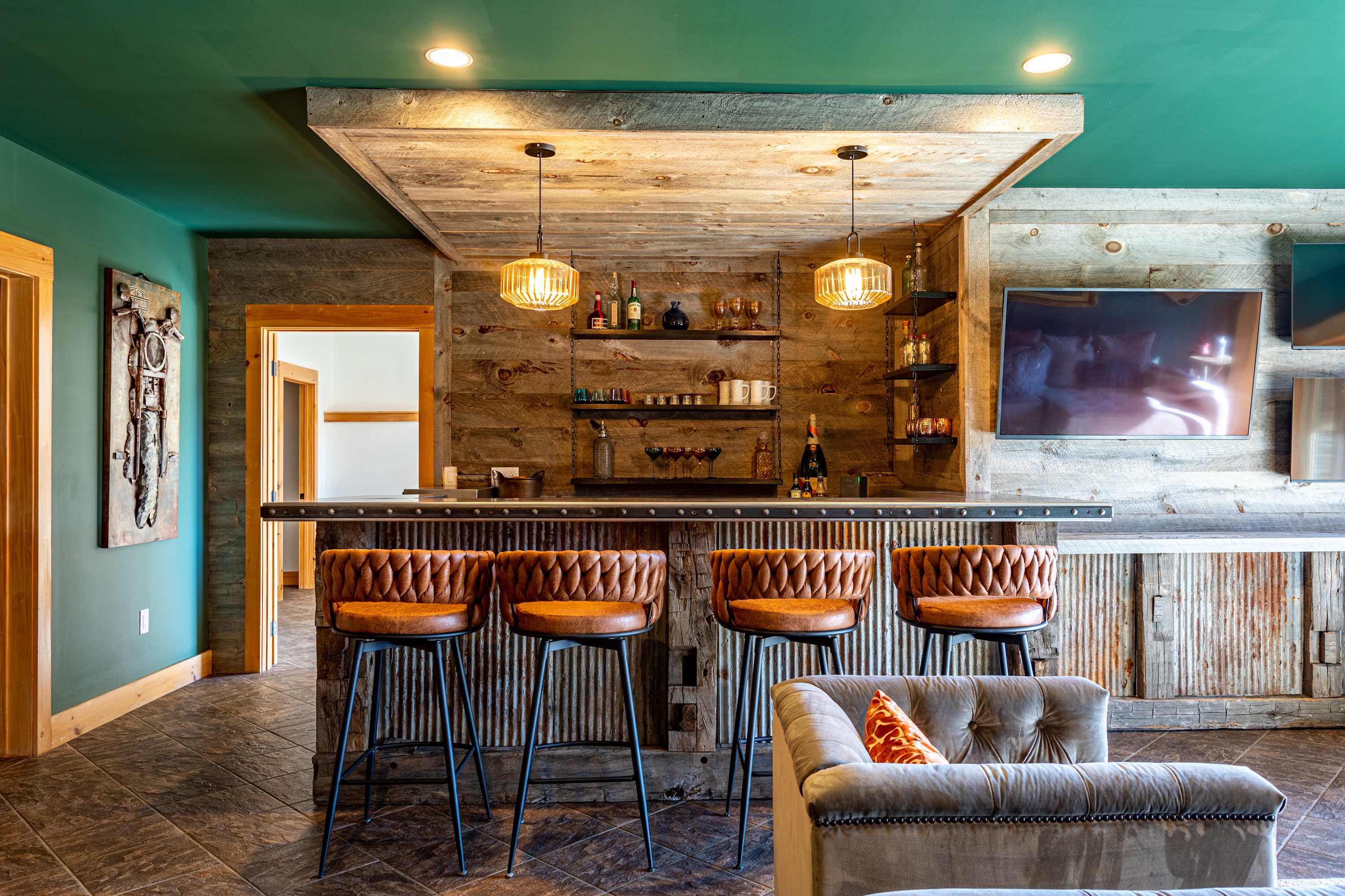 A rustic bar area with a corrugated metal and reclaimed wood facade, topped with a metal counter, and featuring four brown leather bar stools. The back wall is clad in reclaimed wood with open shelving displaying bottles and glassware, all under a green ceiling.