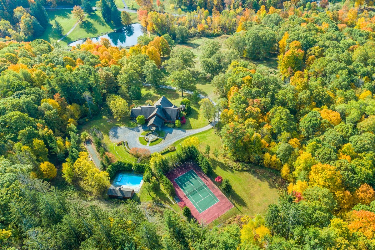 High aerial view of the property showcasing a large house, swimming pool, tennis court, and a pond, all nestled within a vibrant autumn forest with a mix of green, yellow, and orange trees.