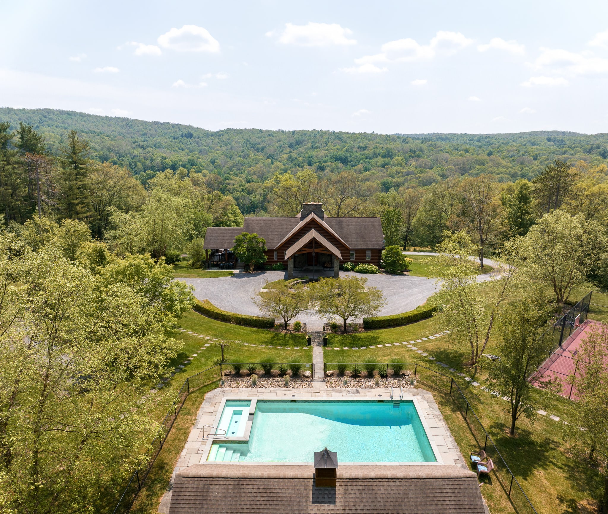 Aerial view of a large log cabin-style house with a circular driveway, surrounded by lush green trees, featuring a rectangular swimming pool in the foreground and distant mountains under a clear sky.