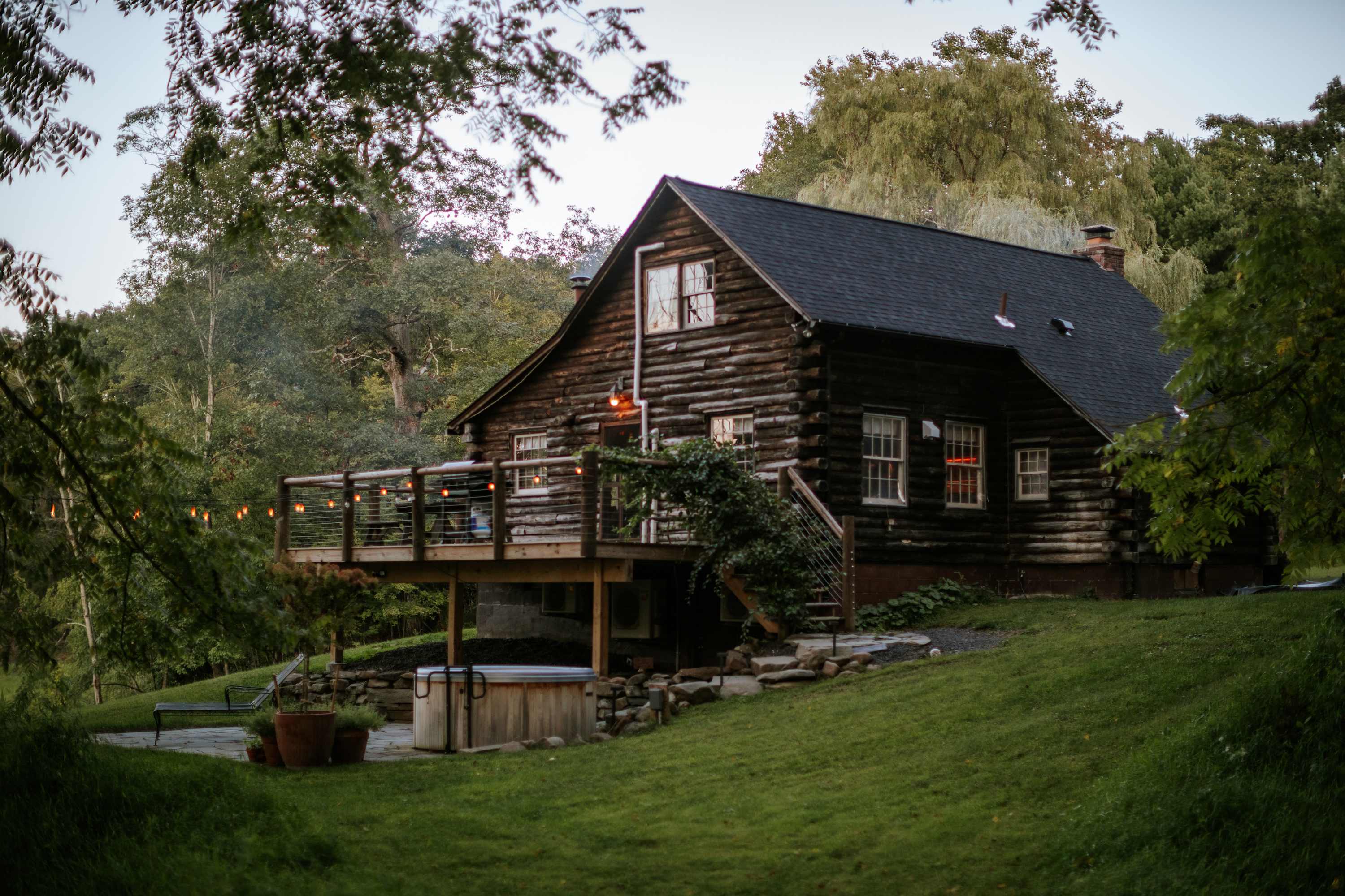 Rear view of the log cabin showcasing a spacious wooden deck with string lights, a hot tub on a stone patio below, and a lush green hillside with dense trees in the background.