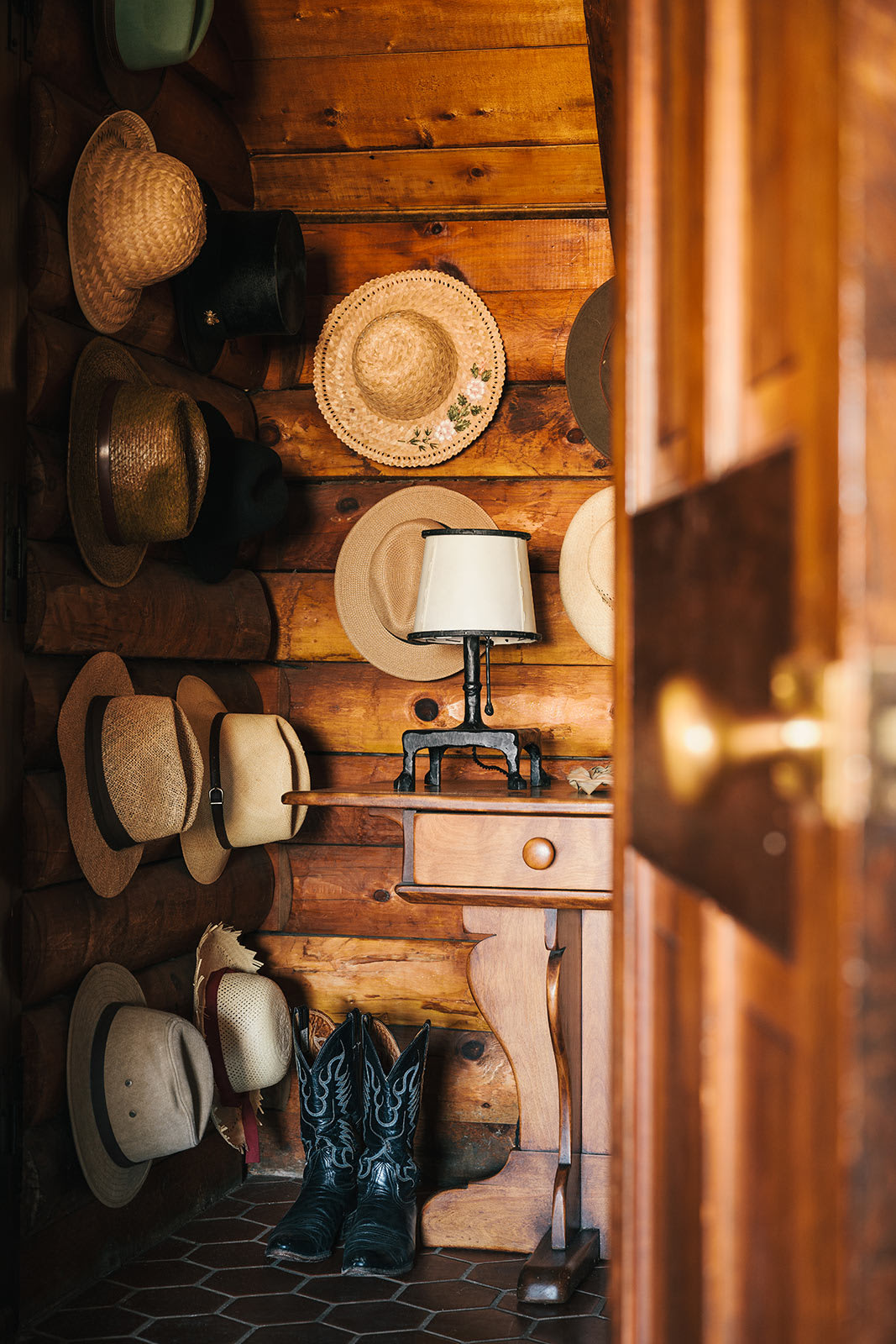 A charming log cabin entryway showcases a collection of straw hats and cowboy boots, creating a rustic welcome. A small wooden console table with a classic lamp adds a touch of warmth to the textured log walls.