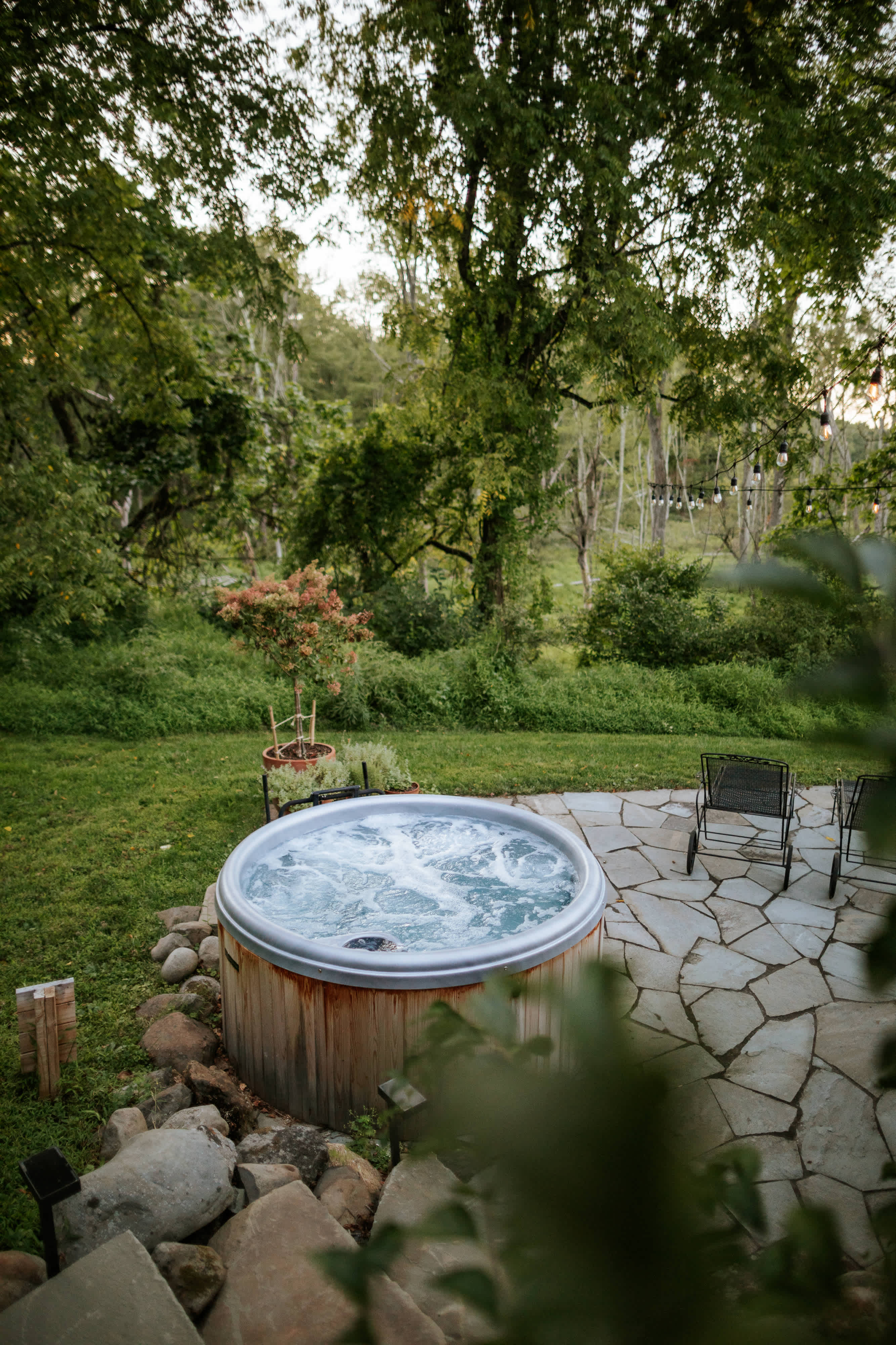 A closer view of the round wooden hot tub on a flagstone patio, showcasing the inviting bubbling water. The natural landscape with mature trees and green grass provides a serene backdrop for relaxation.