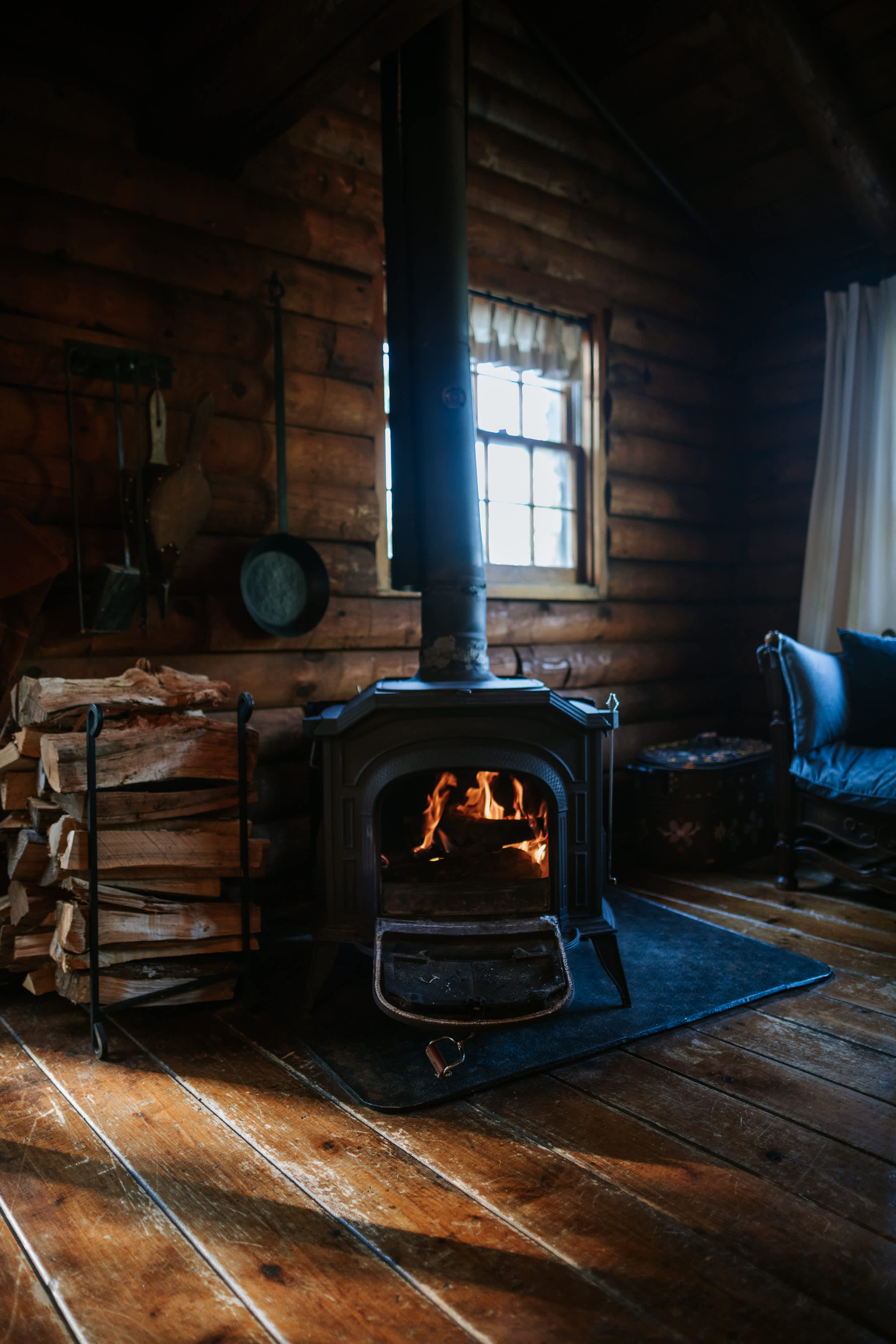 A cozy log cabin living room features a black wood-burning stove with a crackling fire, casting a warm glow on the rustic wooden floors and log walls. A neat stack of firewood sits beside the stove, and a small window with a simple white curtain offers a glimpse outside.