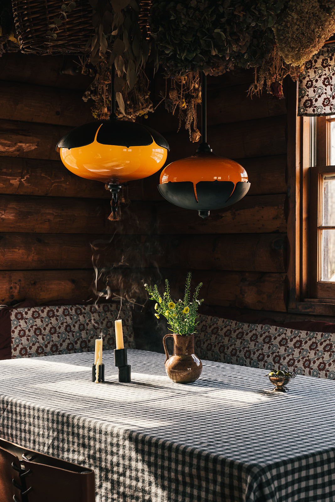 A cozy dining nook inside a log cabin, featuring a checkered tablecloth, a rustic pitcher with flowers, candles, and two unique orange and black pendant lights hanging above.
