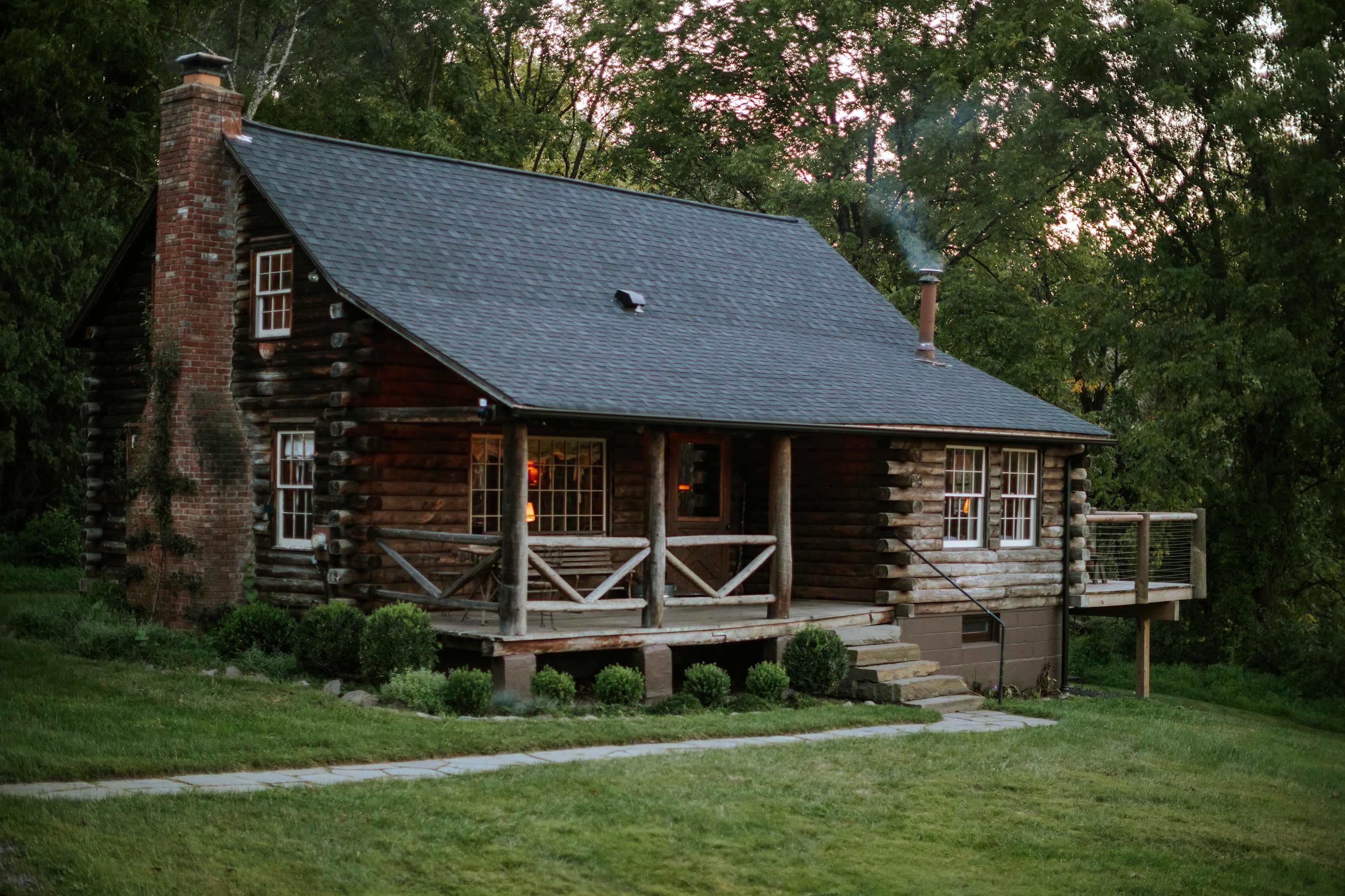 A rustic log cabin with a dark gray shingled roof, a prominent brick chimney, and a covered front porch with wooden railings, nestled in a lush green landscape with trees in the background.