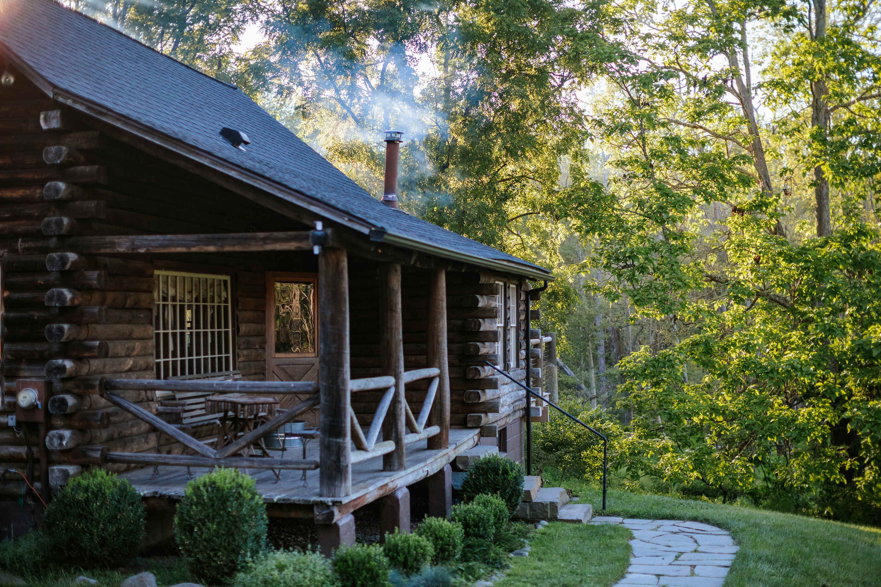 The inviting front porch of a rustic log cabin, featuring a wooden railing, a small dining set, and a stone pathway leading through manicured bushes, surrounded by lush green trees.