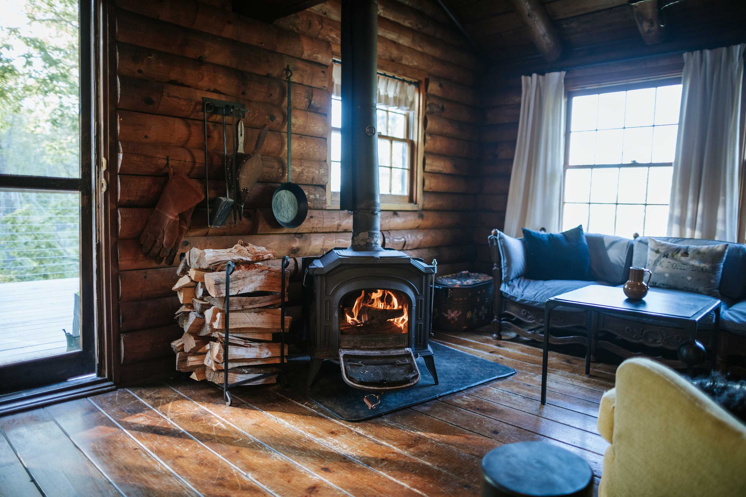 Inviting living space with log walls and hardwood floors, featuring a black wood-burning stove with stacked firewood, a dark blue sofa, and large windows offering views of the outdoors.