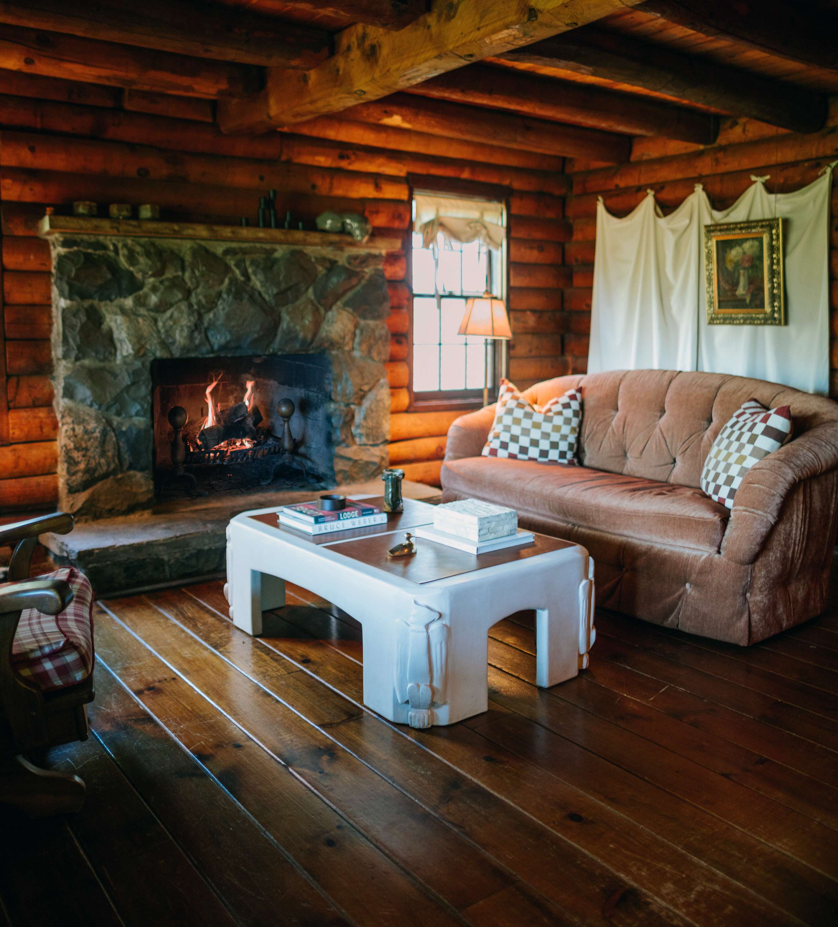 Warm living room with log walls and ceiling beams, centered around a large stone fireplace, a plush brown sofa with patterned pillows, and a white coffee table on polished hardwood floors.