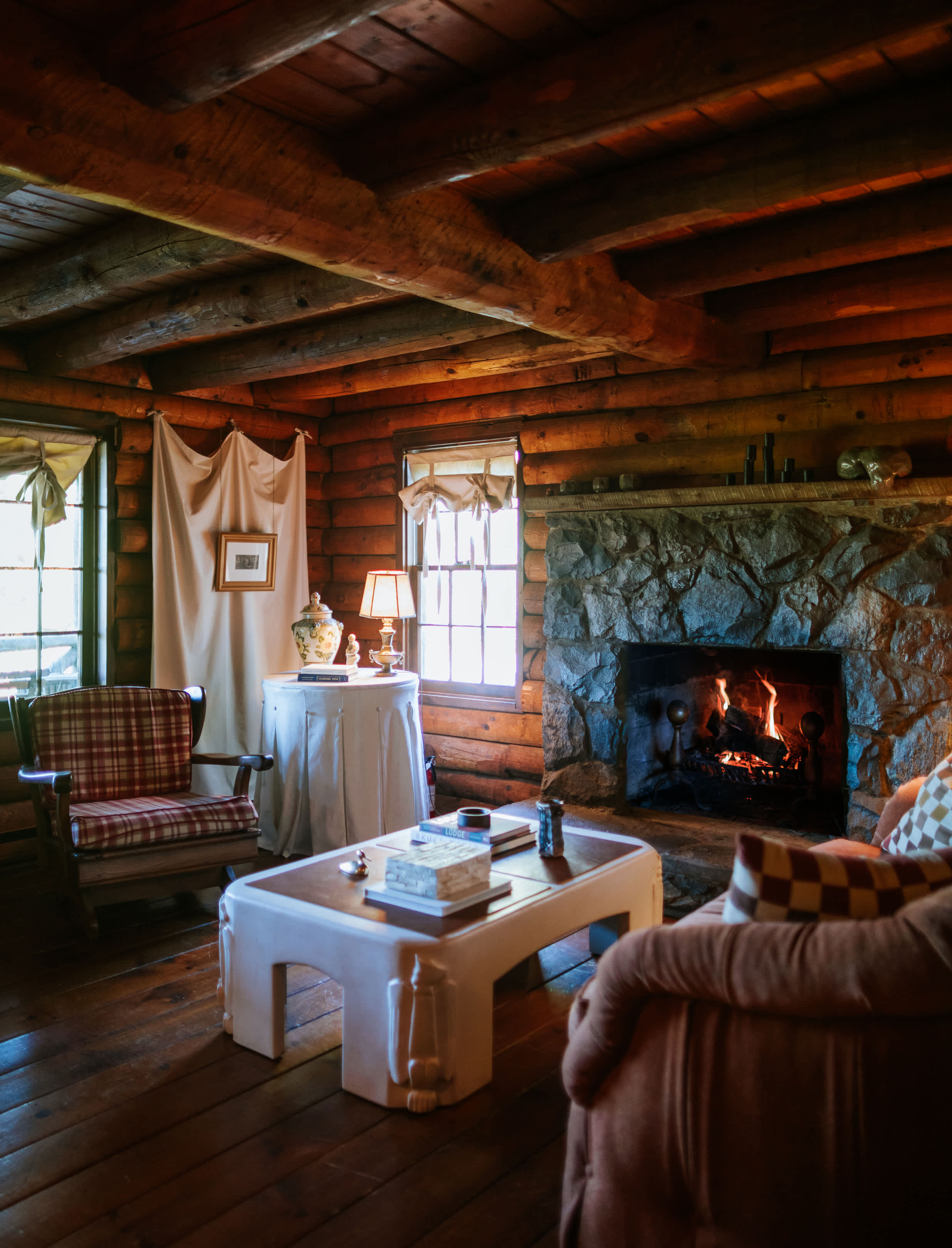 Cozy living room with exposed log walls and ceiling beams, featuring a large stone fireplace with a roaring fire, a plaid armchair, and a white coffee table on dark hardwood floors.