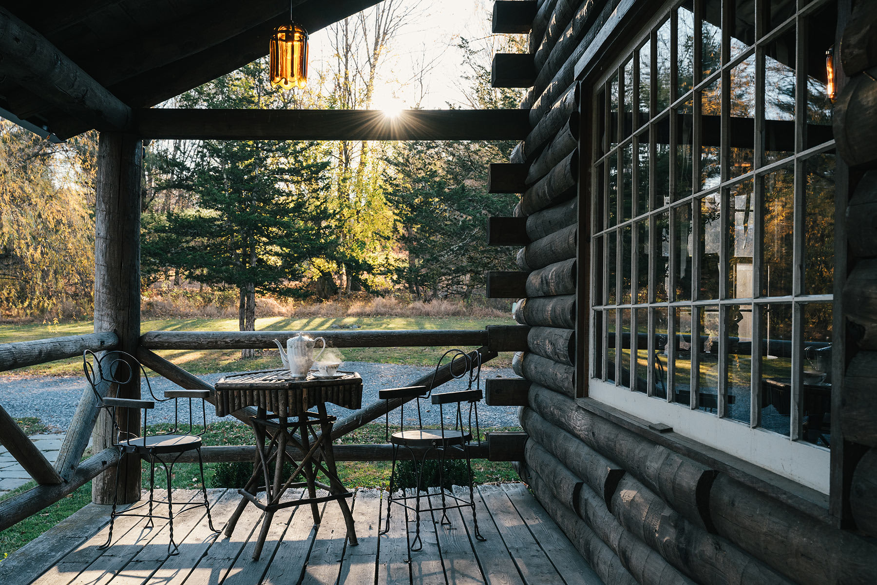 An inviting log cabin porch features a small bistro table and chairs, perfect for enjoying the outdoors. The setting sun casts a warm glow through the surrounding trees, highlighting the rustic log exterior and the charming outdoor lighting.