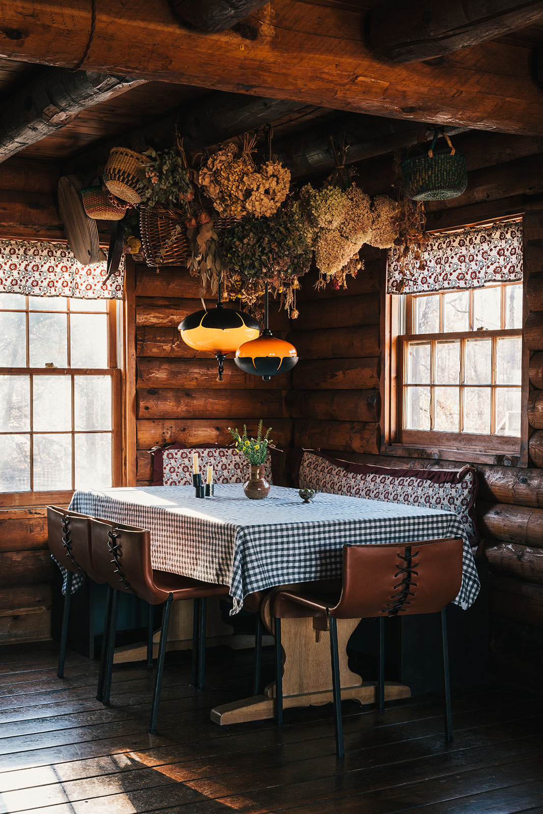 A wider view of the rustic log cabin dining space highlights a table set with a checkered cloth and brown leather chairs, beneath two unique orange and black pendant lights. Sunlight bathes the room, revealing the textured log walls and the array of dried botanicals suspended from the ceiling.