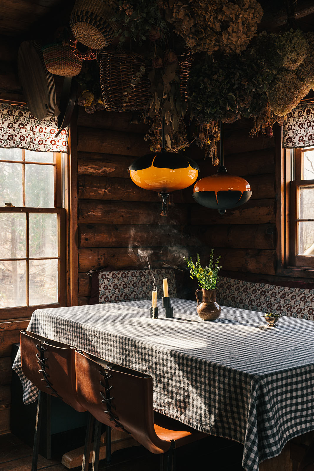 A cozy log cabin dining area features a table draped with a black and white checkered cloth, surrounded by brown leather chairs, and illuminated by two distinctive orange and black pendant lights. Dried botanicals hang from the rustic wooden ceiling, and sunlight streams through windows with patterned valances.