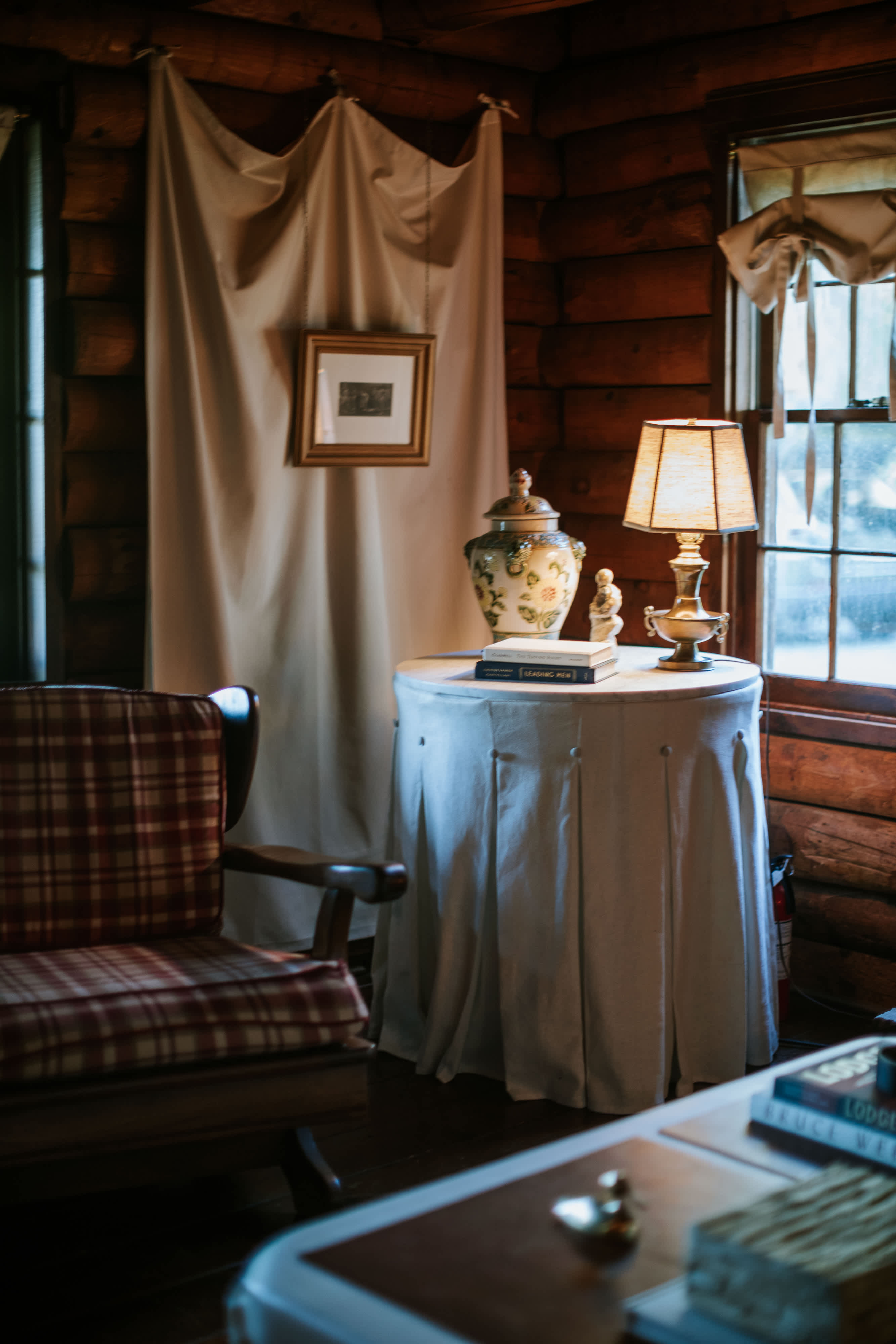 A cozy corner of the log cabin living room features a plaid armchair and a round table draped with a white skirt, holding a classic lamp and a decorative vase. A framed picture hangs on a fabric-draped wall, adding a homely touch to the rustic log interior.