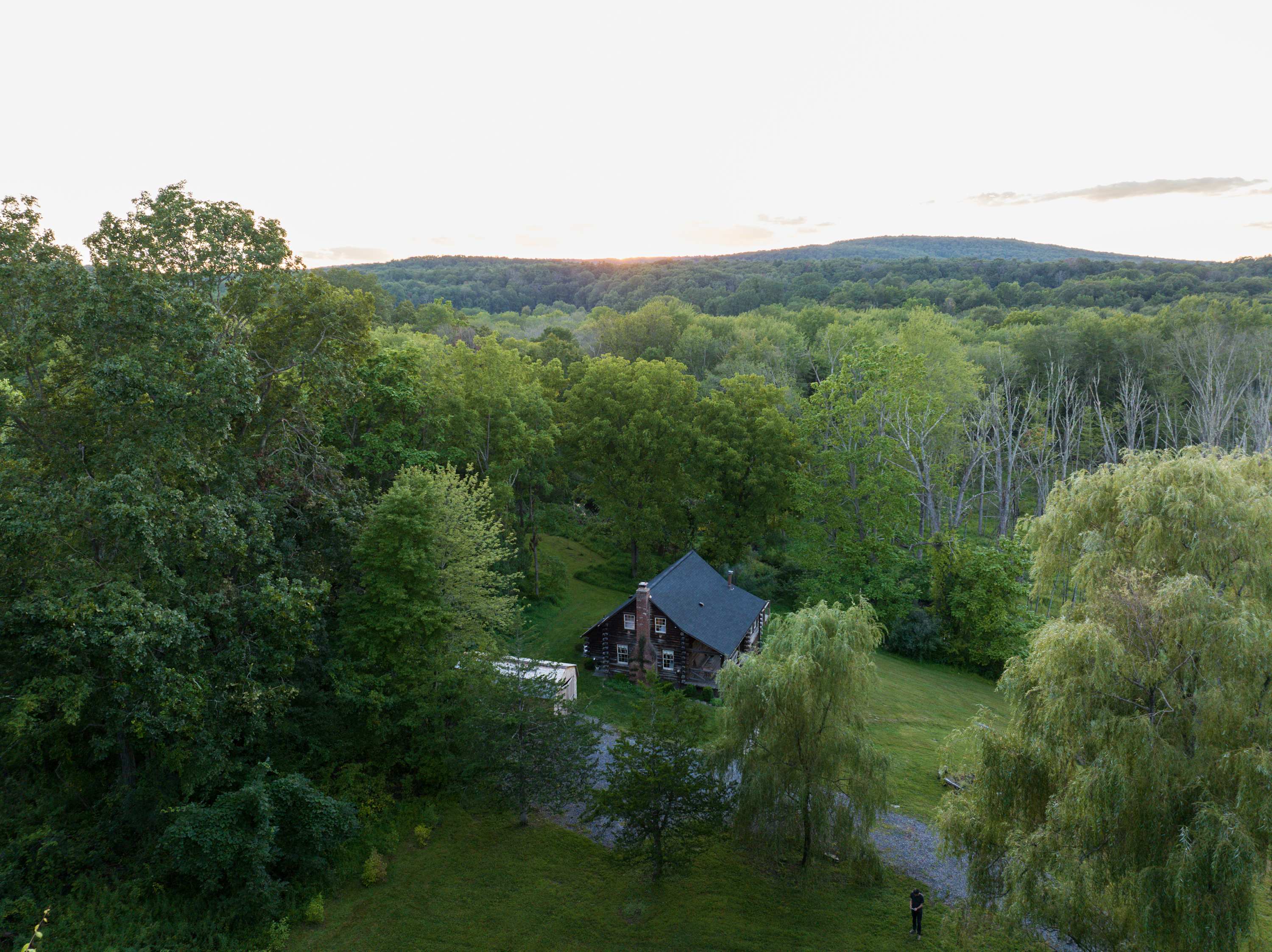 An aerial view of a charming log cabin with a dark roof, nestled in a clearing surrounded by dense green forests and distant rolling hills under a soft evening sky.