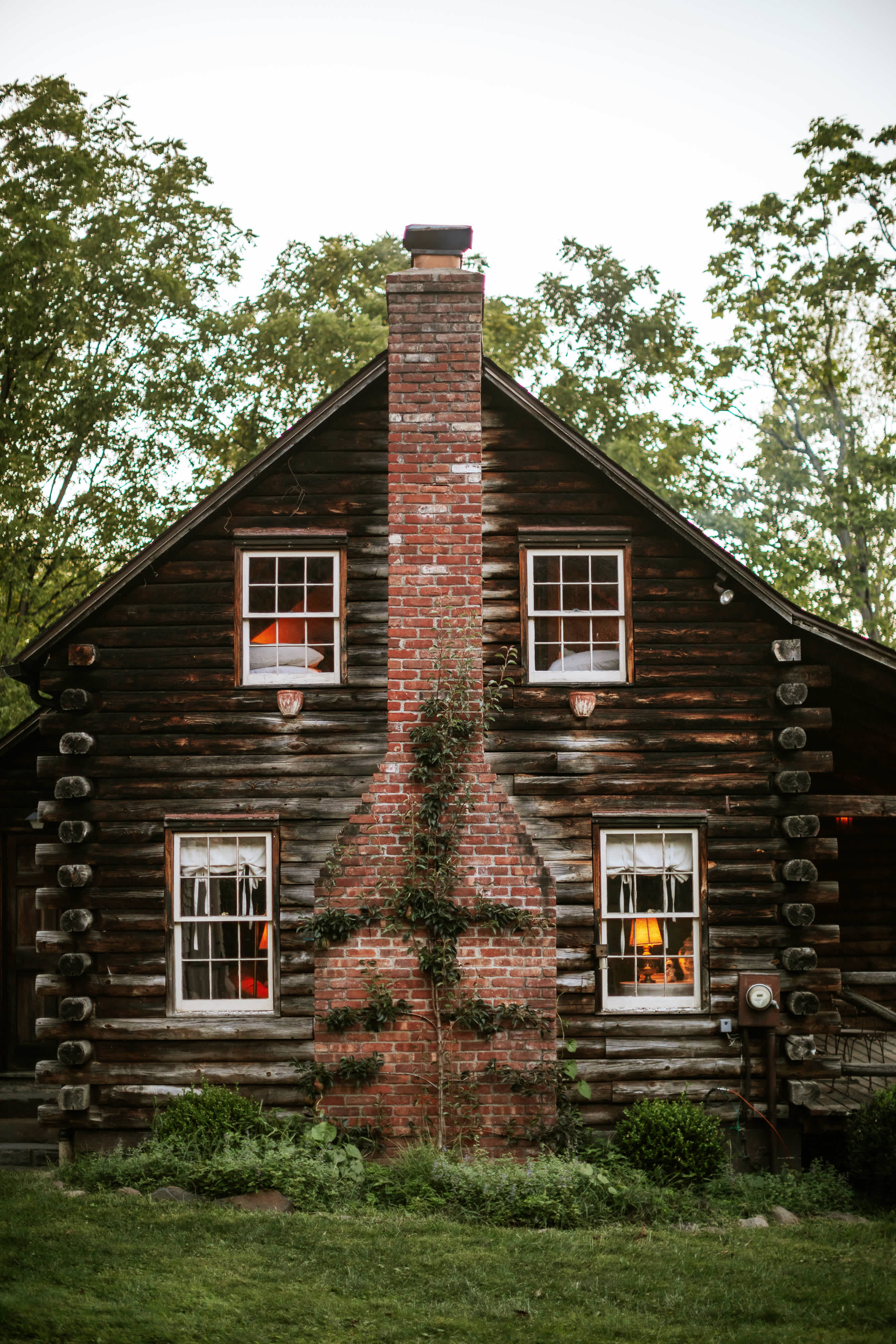 Front facade of a dark log cabin featuring a tall brick chimney covered in ivy, white-framed windows with multiple panes, and a dark wooden door, surrounded by green grass and trees.
