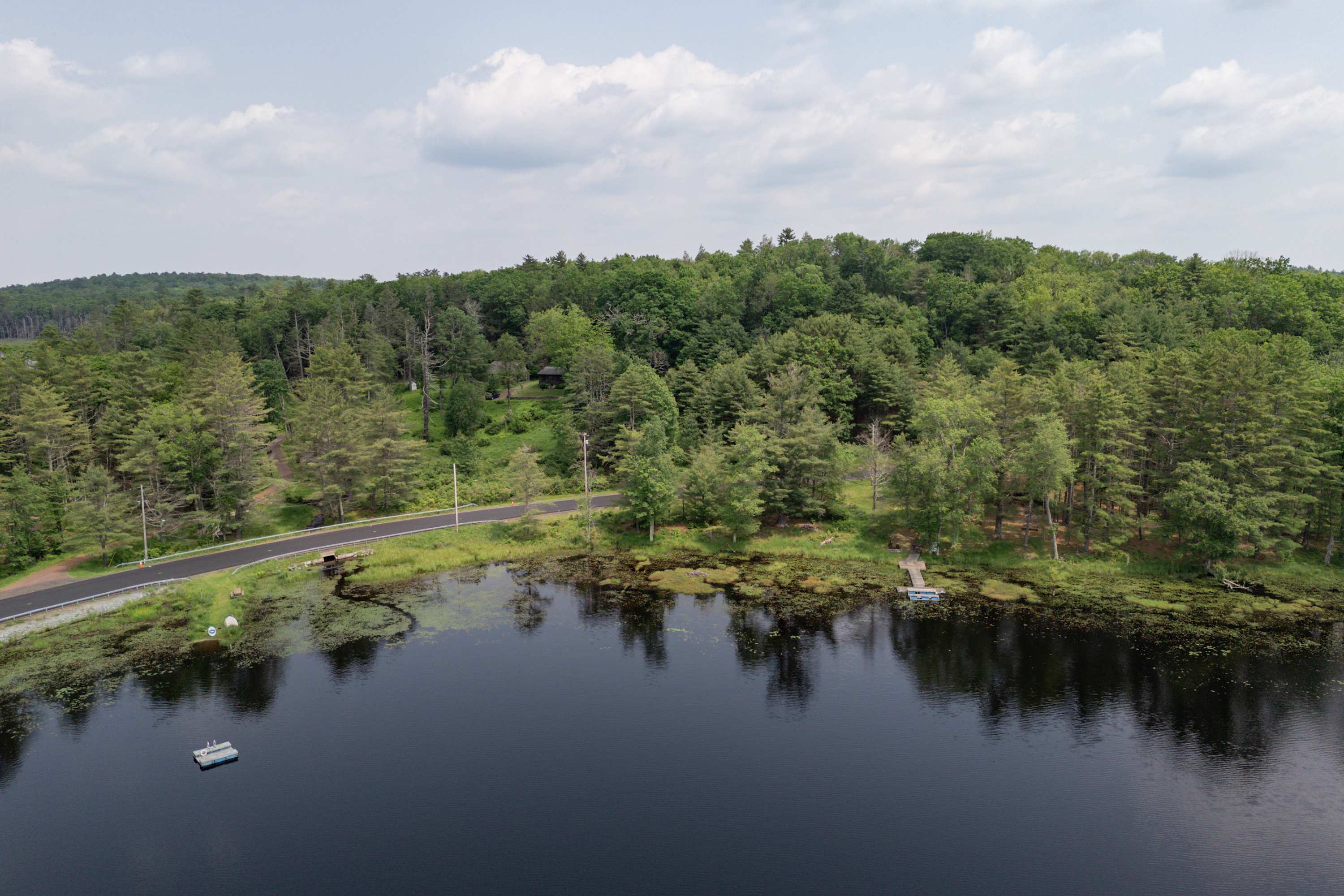 An aerial perspective shows a calm lake with a small dock and a road running alongside, bordered by a lush green forest with a house visible in the distance.