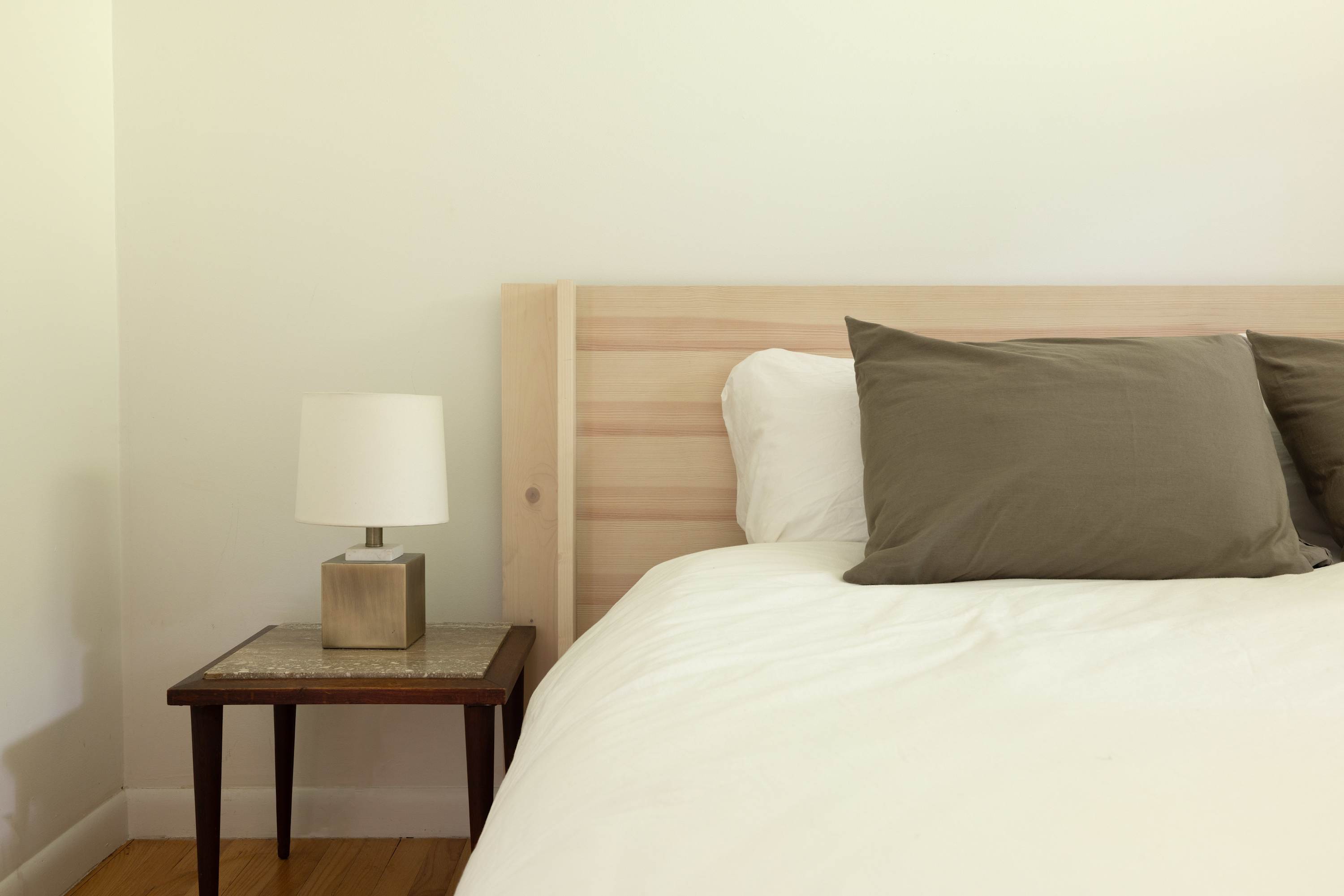 Close-up of a light wood headboard and a vintage-style nightstand with a marble top, holding a modern white lamp. Two grey throw pillows accent the white bedding.