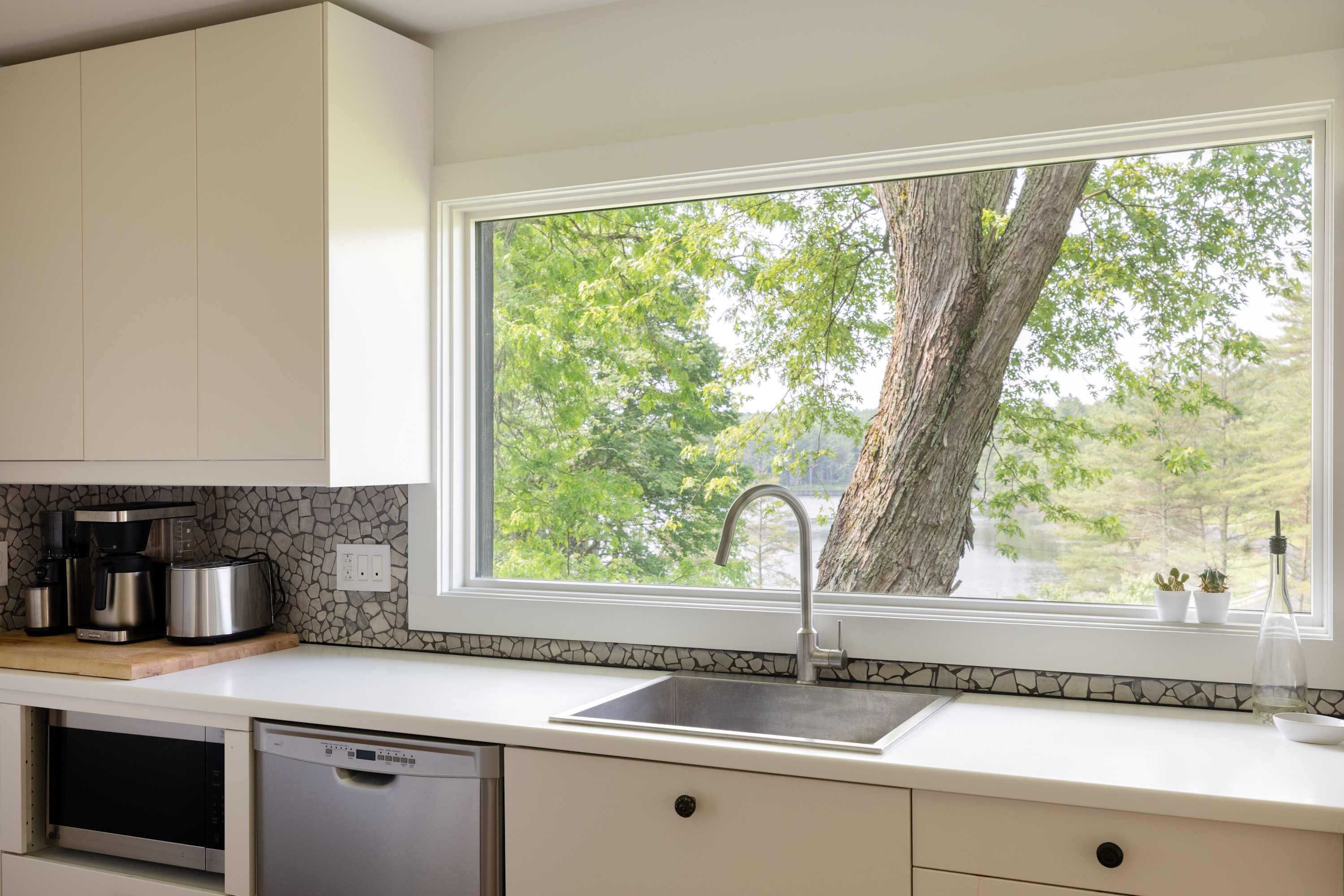 Kitchen counter with a stainless steel sink and modern faucet, flanked by white cabinets and a stone mosaic backsplash. A large window above the sink offers a view of a mature tree and the lake.