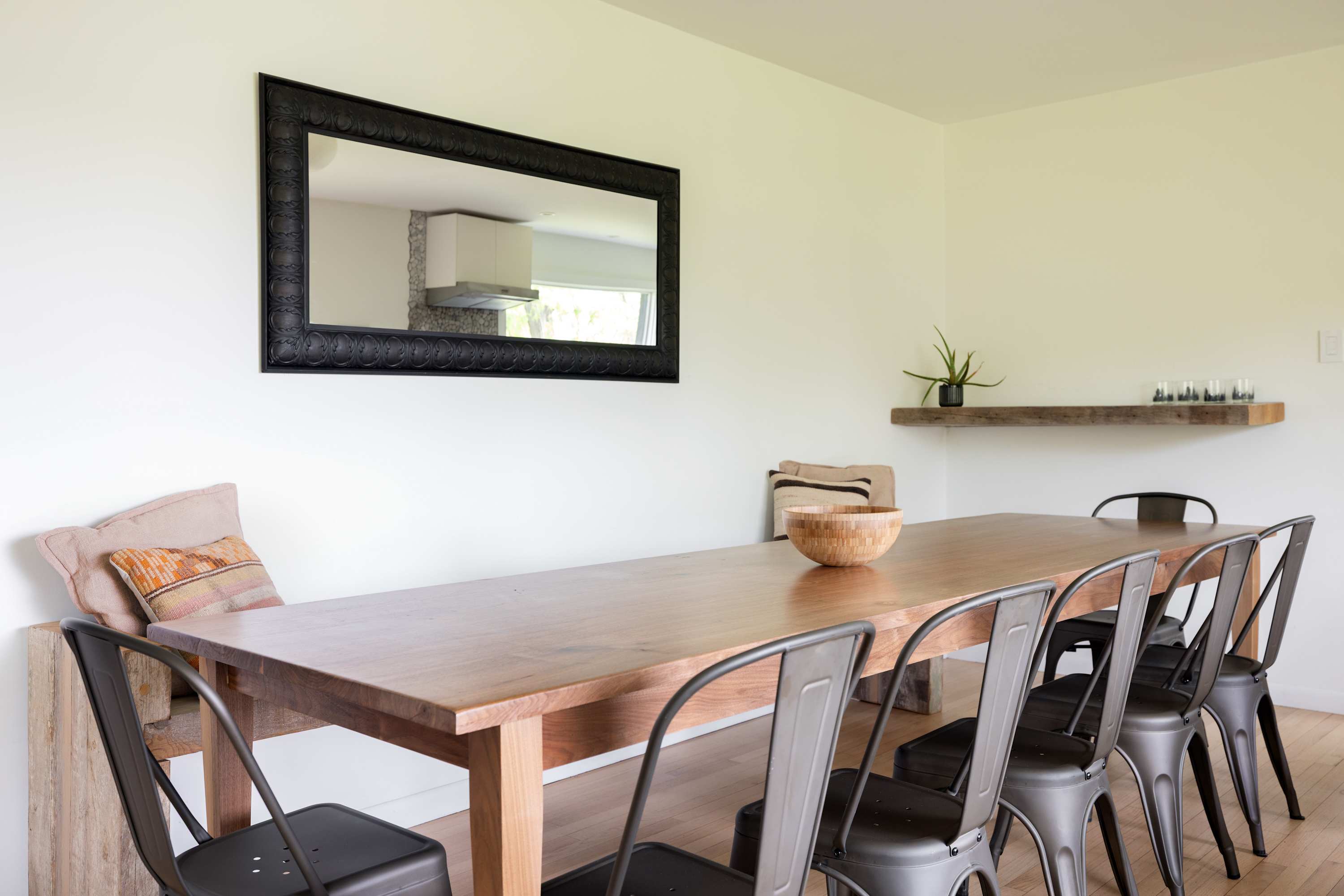 Spacious dining area with a long wooden table, surrounded by black metal industrial-style chairs, and a large black framed mirror reflecting the kitchen. A rustic wooden shelf holds a plant.