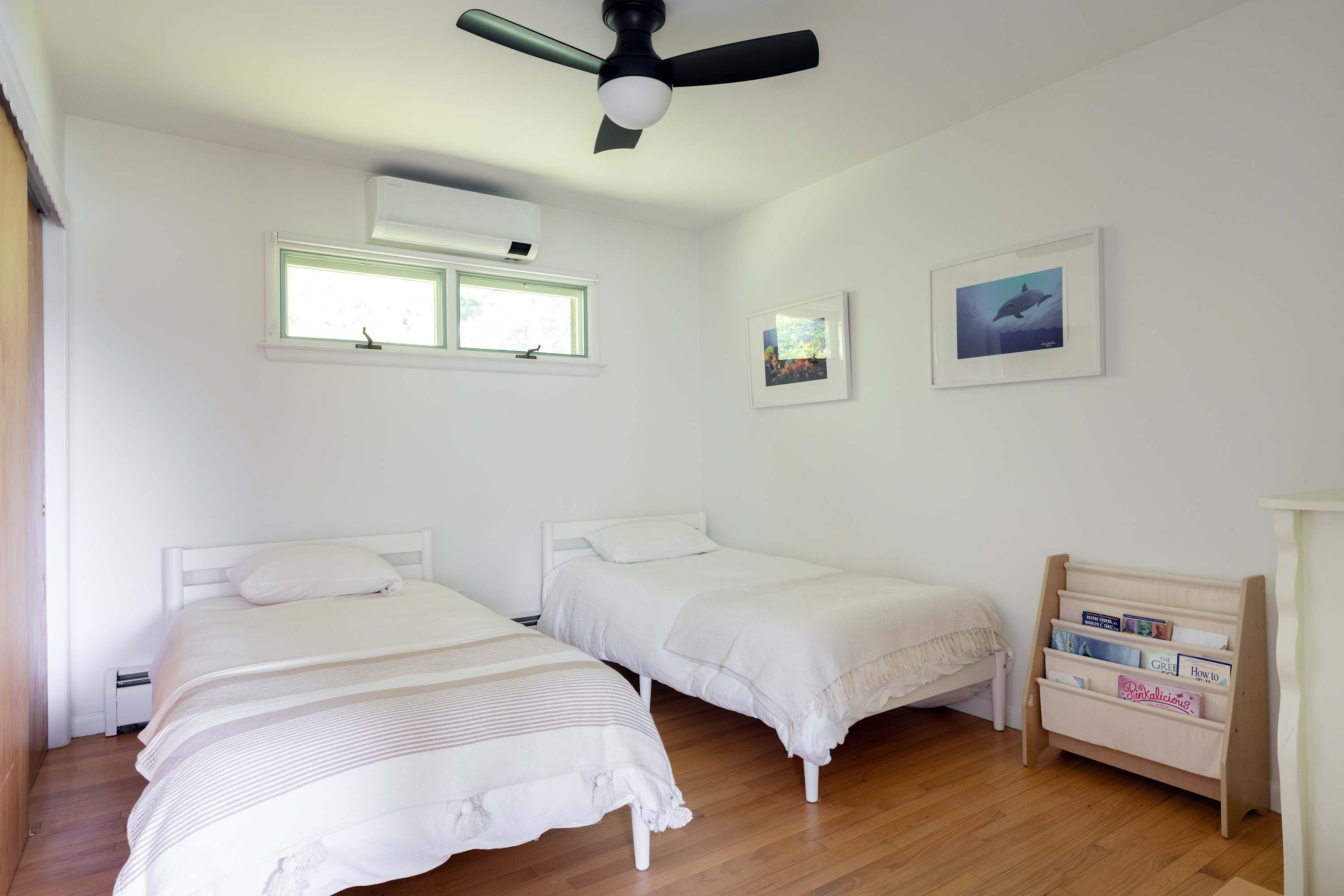 A bright bedroom features two white twin beds with striped and textured white bedding, hardwood floors, and a ceiling fan, with two framed pictures on the white wall.