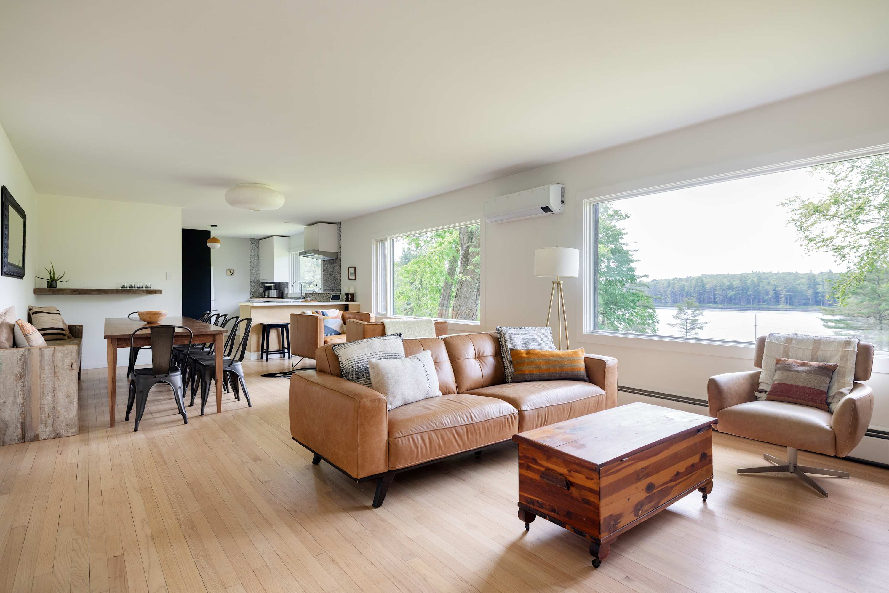 Bright open-concept living space with light hardwood floors, a tan leather sofa, a wooden coffee table, and large windows offering a panoramic lake view. A dining area and kitchen are visible in the background.