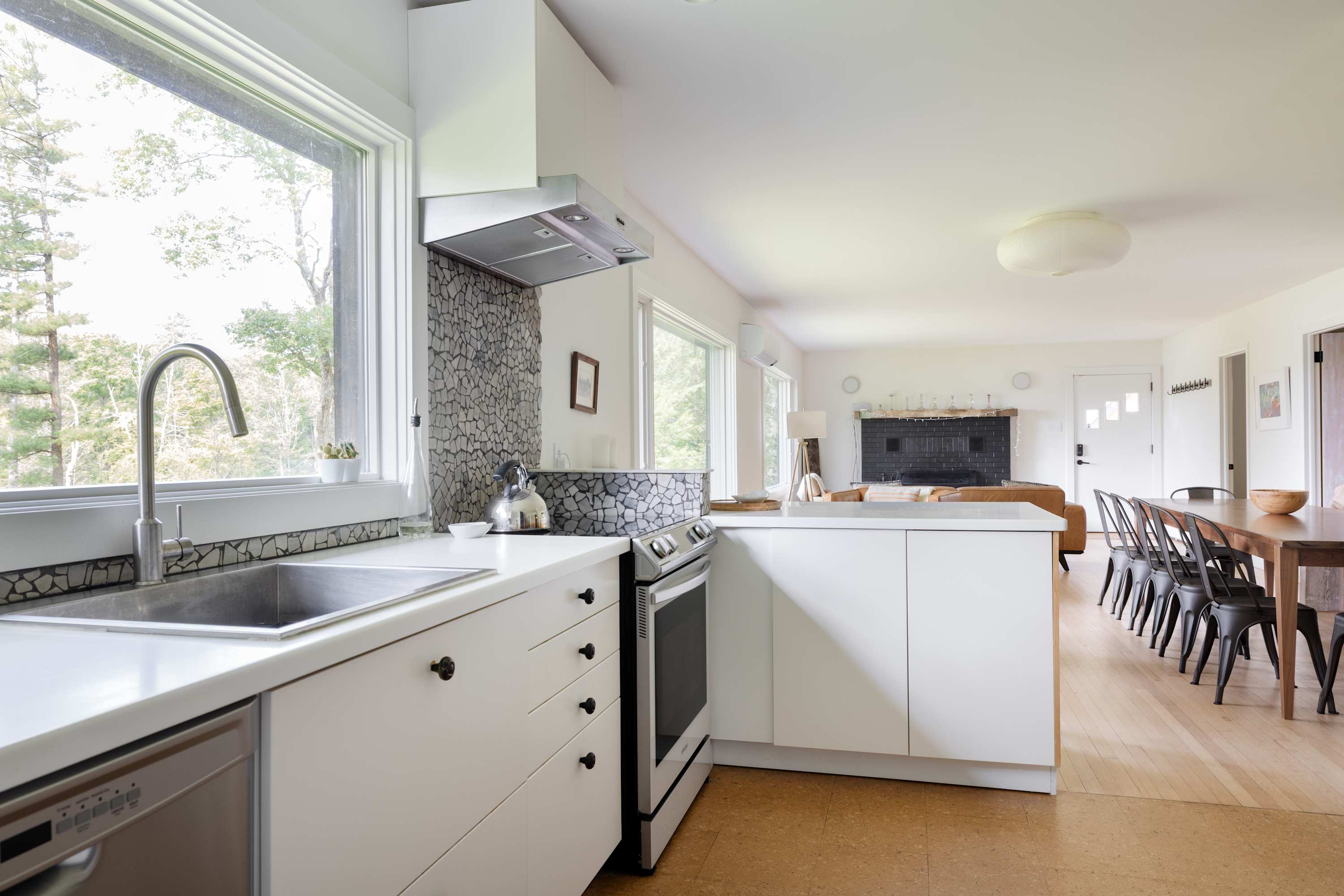 Open-concept kitchen featuring white cabinetry, a stainless steel sink, and a stove with a range hood, all set against a stone mosaic backsplash. The space flows into the dining and living areas.
