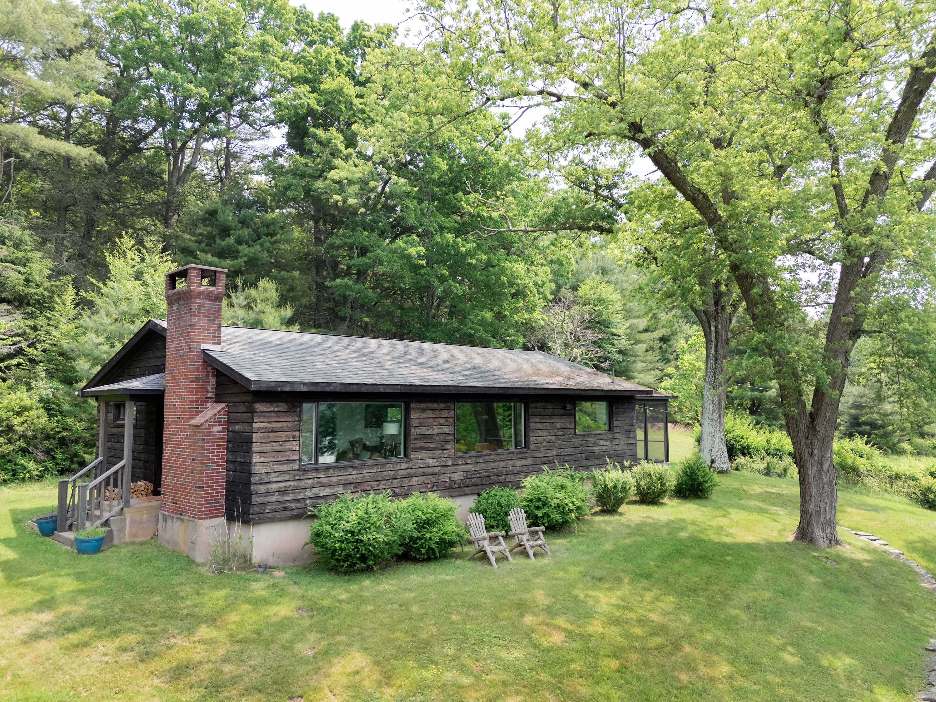 The rustic wooden house features a prominent brick chimney and large windows, surrounded by a well-maintained green lawn and mature trees under a bright sky.