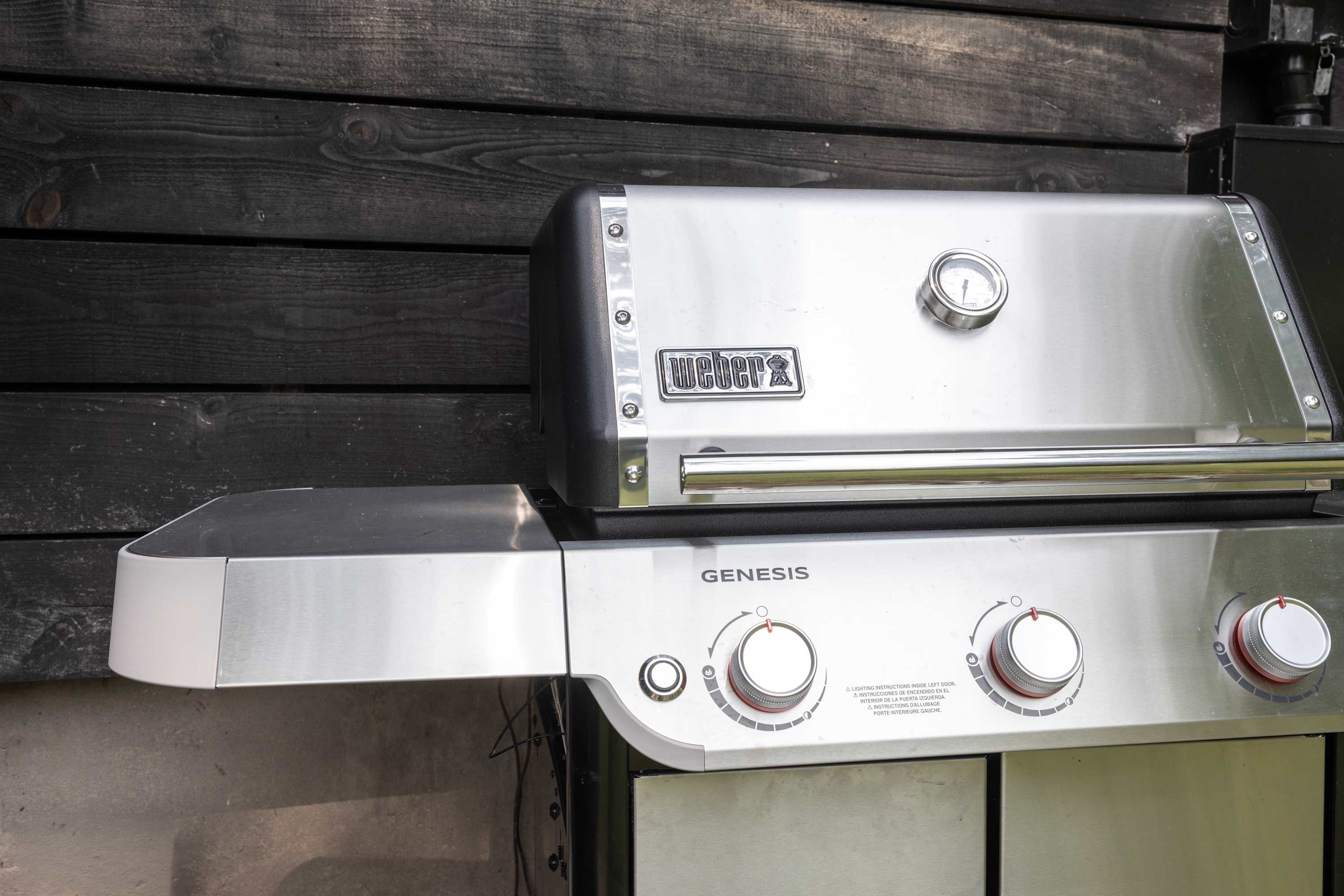 A shiny stainless steel Weber Genesis gas grill with control knobs and a temperature gauge is positioned against a dark wooden wall.