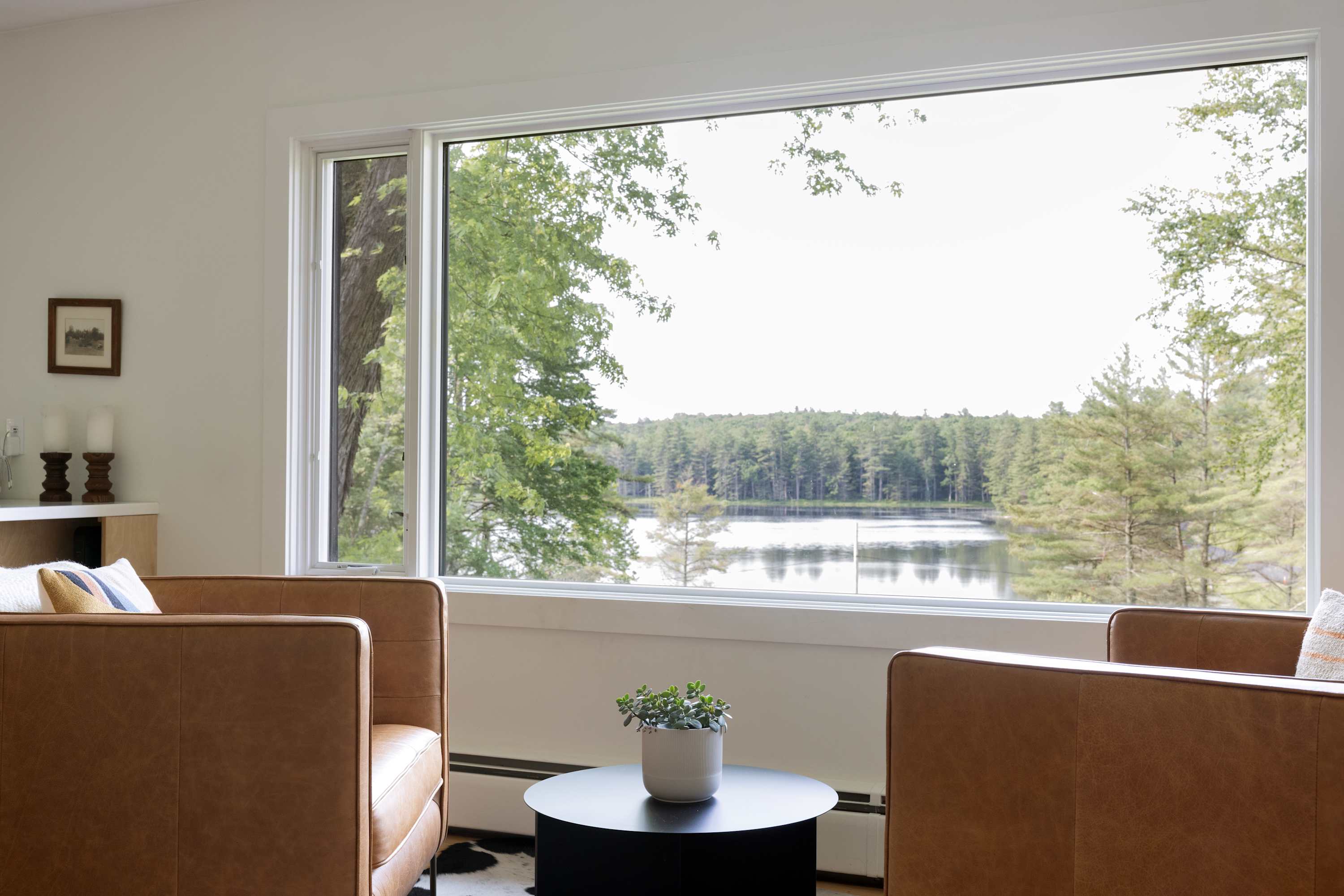 Bright living room featuring two tan leather armchairs, a round black coffee table with a plant, and a large picture window offering expansive views of a serene lake and surrounding forest.