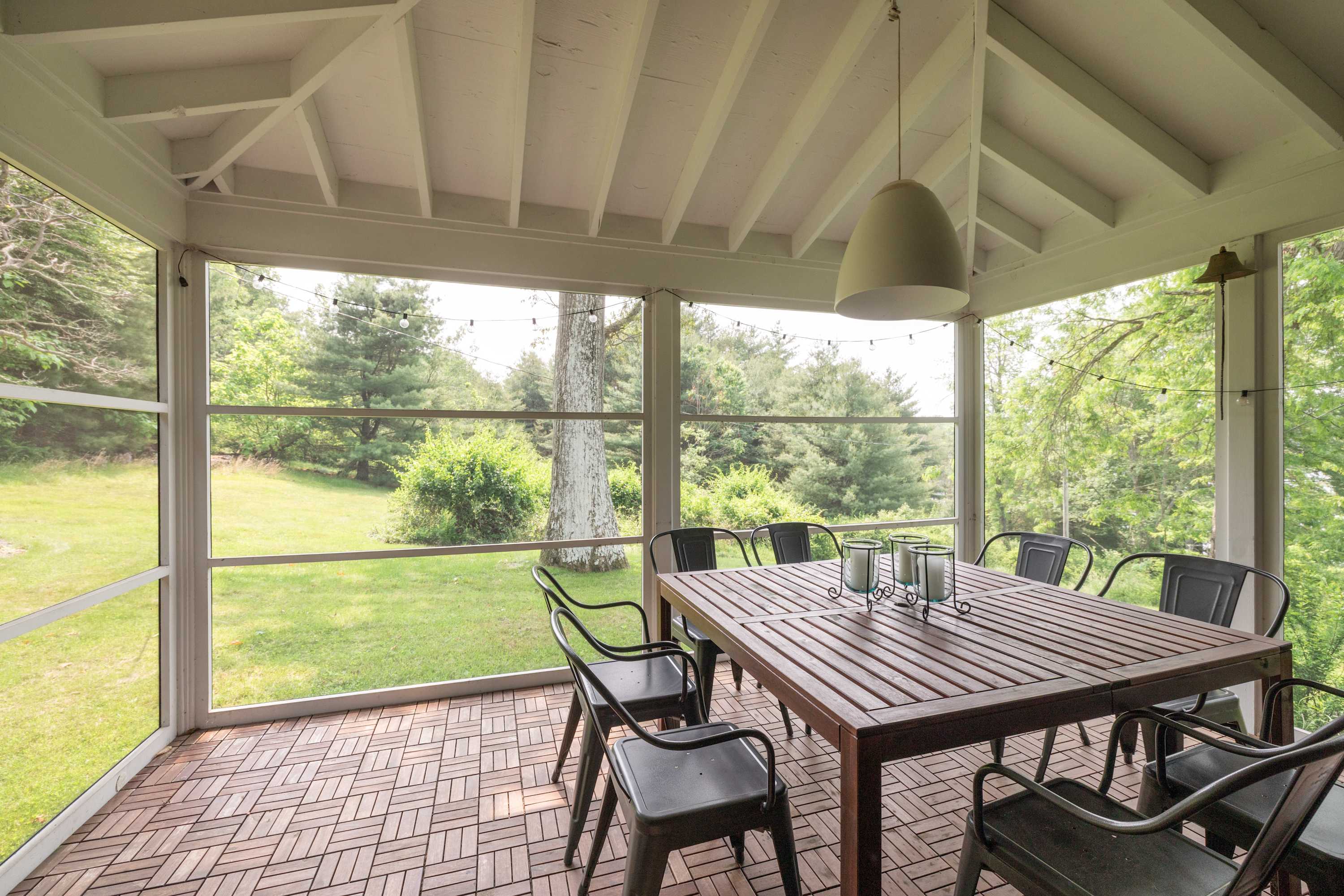 Screened-in porch with a dark wooden dining table and black metal chairs, featuring a light-colored pendant lamp and views of the surrounding green yard and trees.