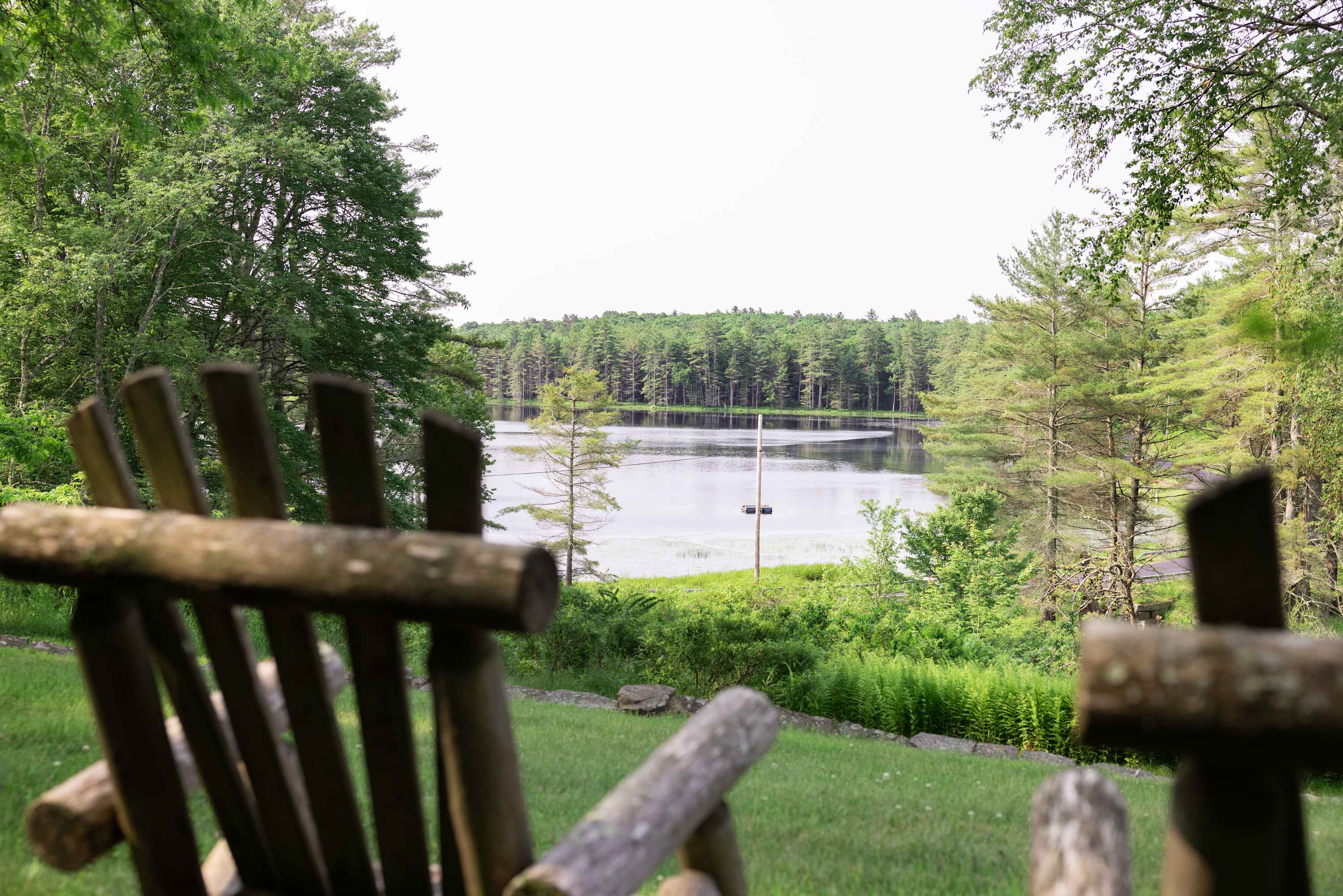 Scenic outdoor view from a grassy yard, featuring a rustic wooden Adirondack chair in the foreground and a wide expanse of a tranquil lake surrounded by dense green forests in the distance.