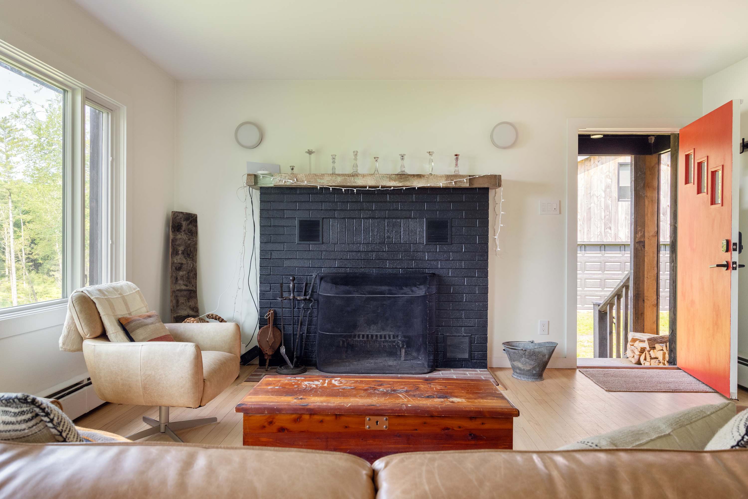 Cozy living room featuring a black brick fireplace with a rustic wooden mantel, light hardwood floors, a comfortable armchair, and a vibrant red front door leading to the exterior.