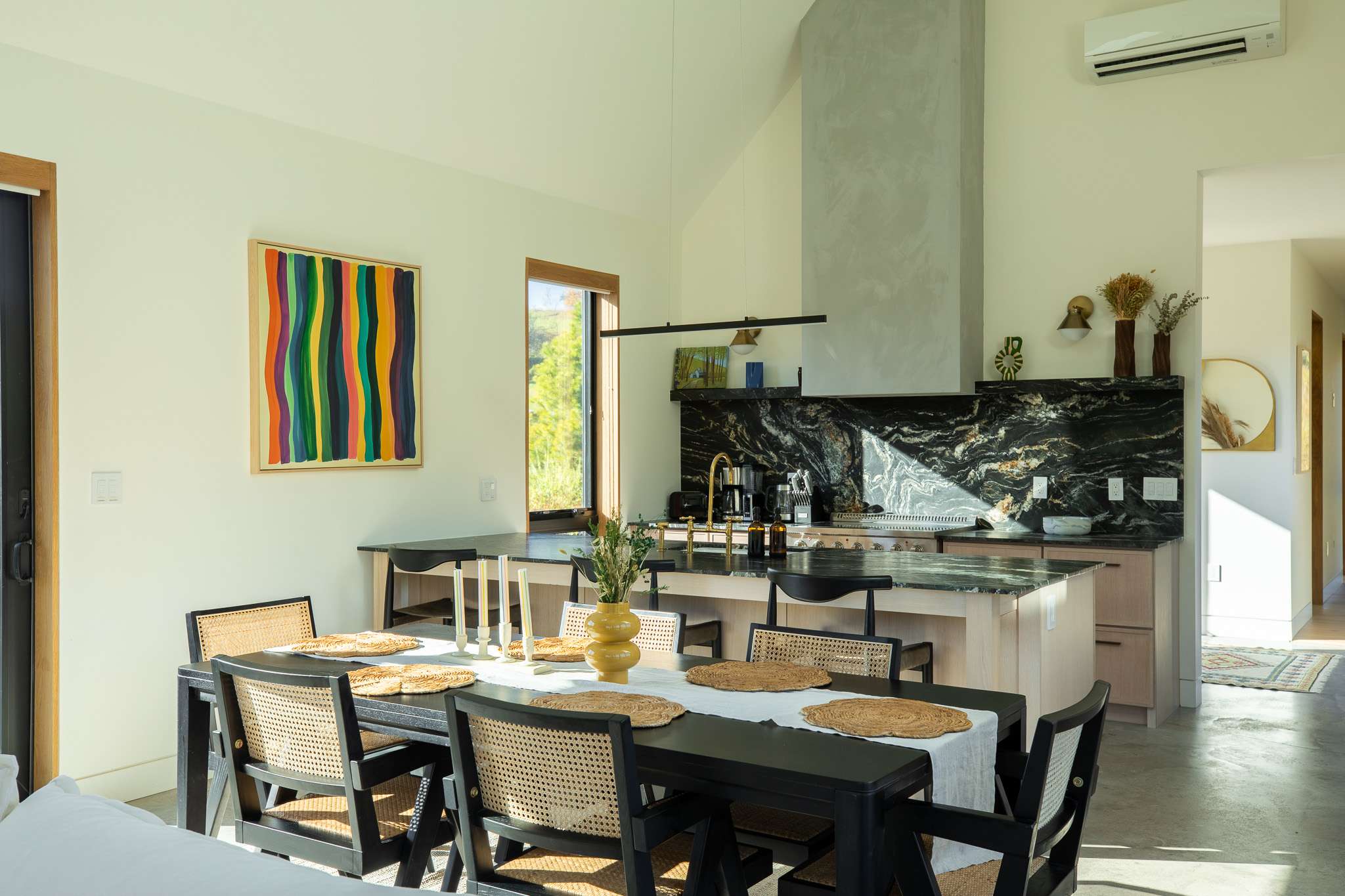 The dining area features a black rectangular table with six black chairs, each with a cane back, set on a light rug. A vibrant striped abstract painting hangs on the wall, and a large window overlooks a scenic landscape, connecting the indoor space with nature.