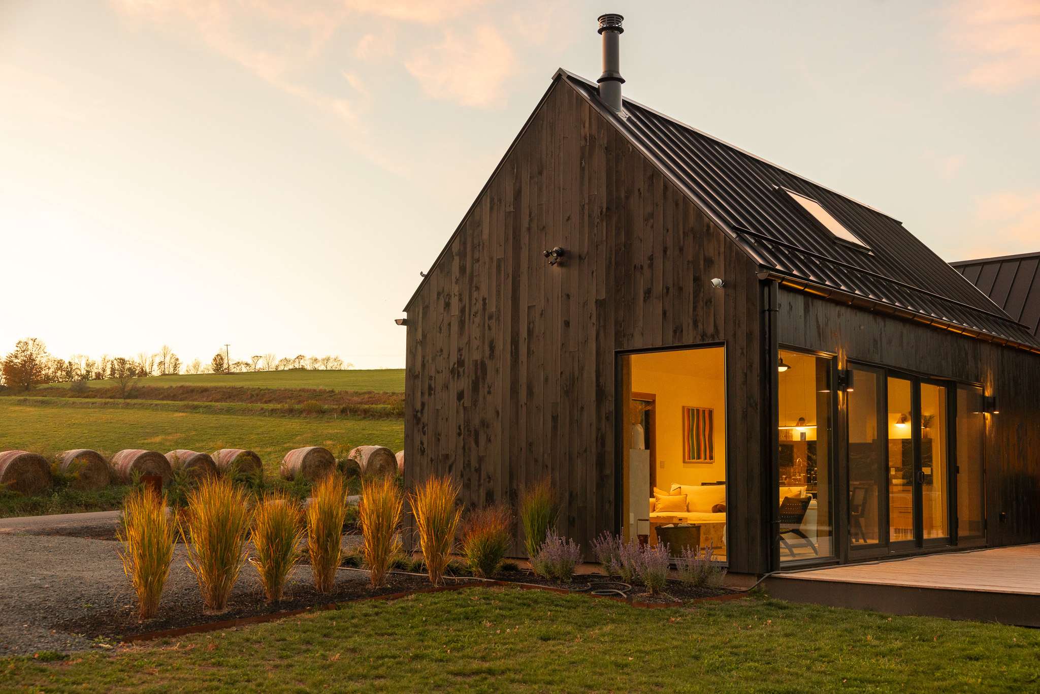 Angled view of the black modern barn-style house with a dark metal roof, large glass doors, and a chimney. The house is set against a golden sunset sky, with hay bales in the foreground and rolling hills in the distance.