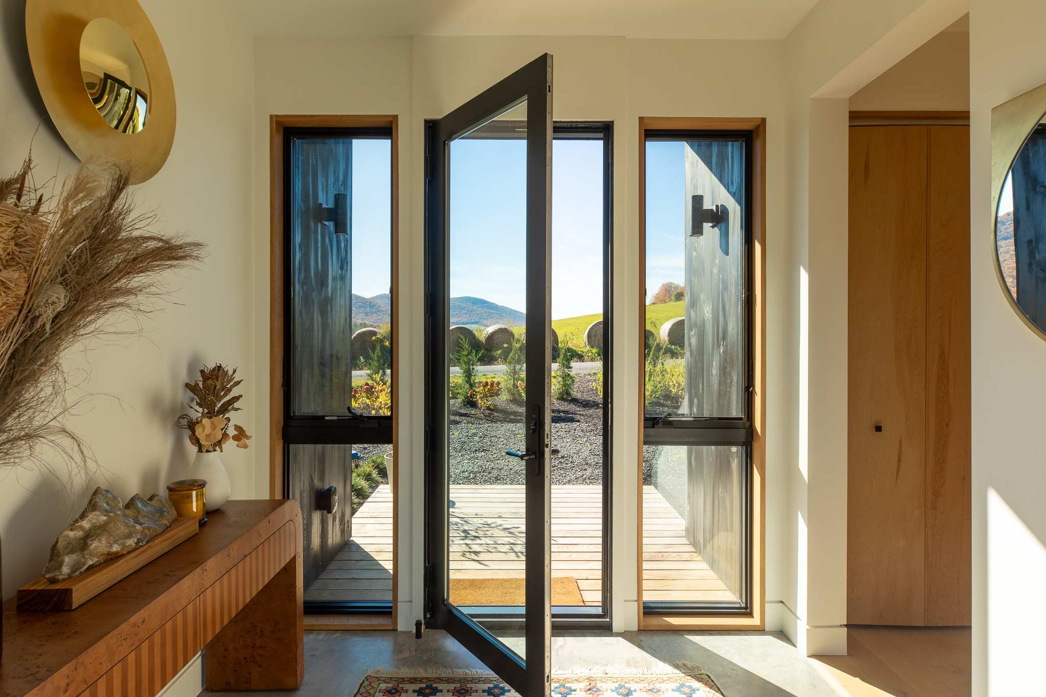Bright and inviting foyer with a modern glass pivot door and side windows, offering a direct view of the outdoor deck and the scenic rolling hills beyond. A wooden console table and abstract art adorn the space.