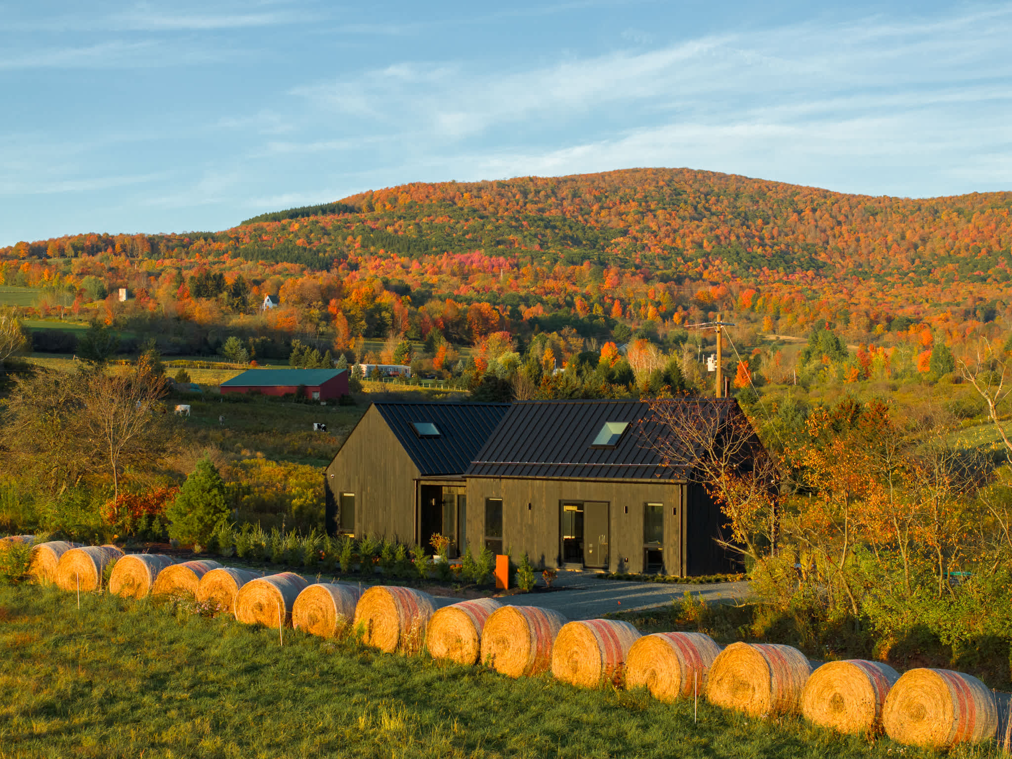 Aerial view of the black modern barn-style house nestled in a vibrant autumn landscape with rolling hills covered in colorful fall foliage. Hay bales line the foreground, and distant farm buildings are visible under a clear sky.