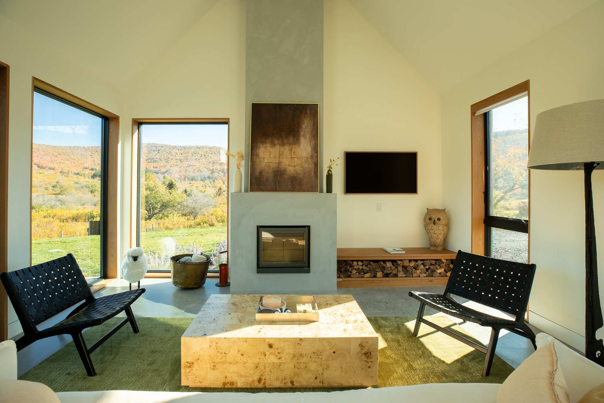 Bright living room with a concrete fireplace, a large square wooden coffee table, two black woven chairs, and expansive windows offering autumn mountain views.